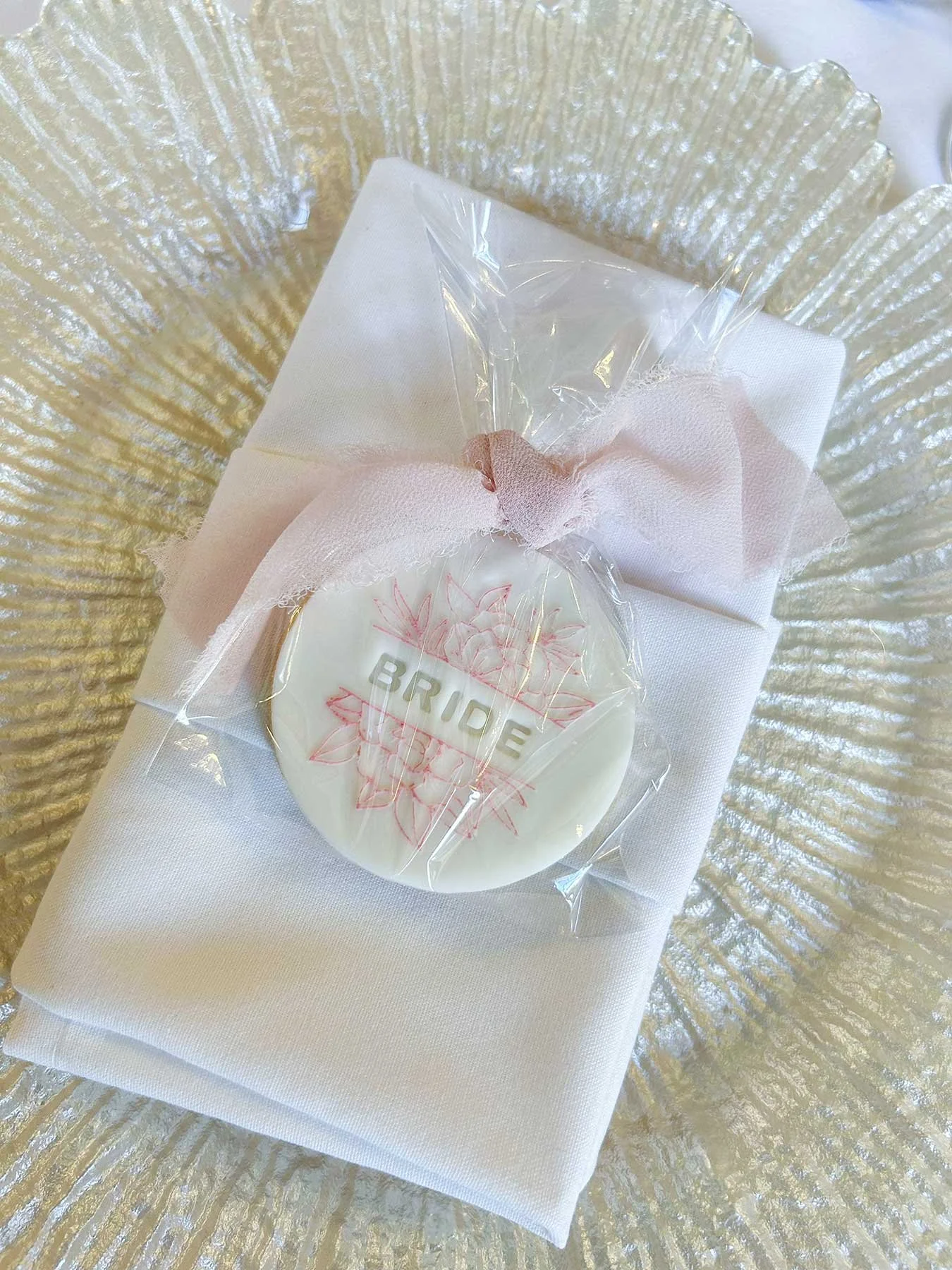 A white napkin with wrapped wedding favor on top, featuring a round white chocolate labeled 'BRIDE' with pink floral designs, placed on a gold-rimmed charger plate.