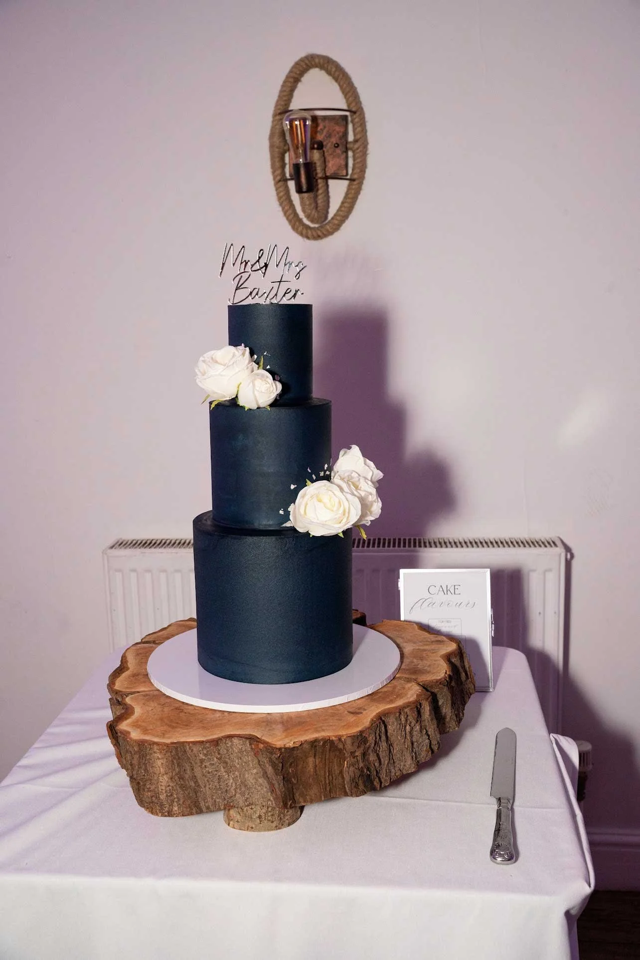 Three-tier navy blue wedding cake with white roses on a wooden slab, topped with a custom wire cake topper reading "Mr & Mrs Baxter."