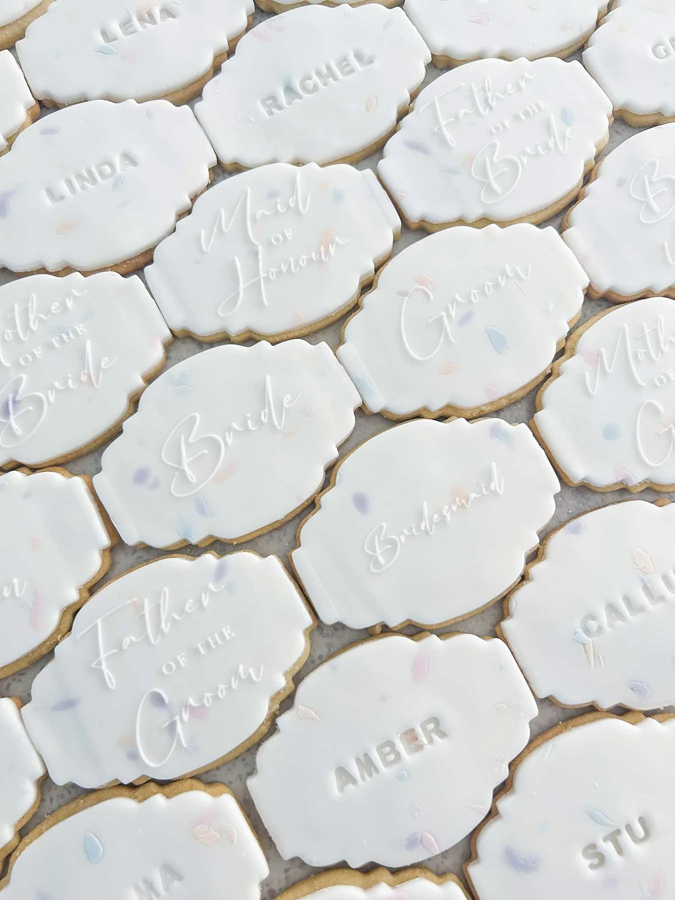 Cookies decorated with white icing and labels for a wedding, including titles like "Mother of the Bride," "Mother of the Groom," "Bride," "Groom," "Mother of the Groom," "Mother of the Bride," "Bridesmaid,"  and individual names such as "Rachel," "Li