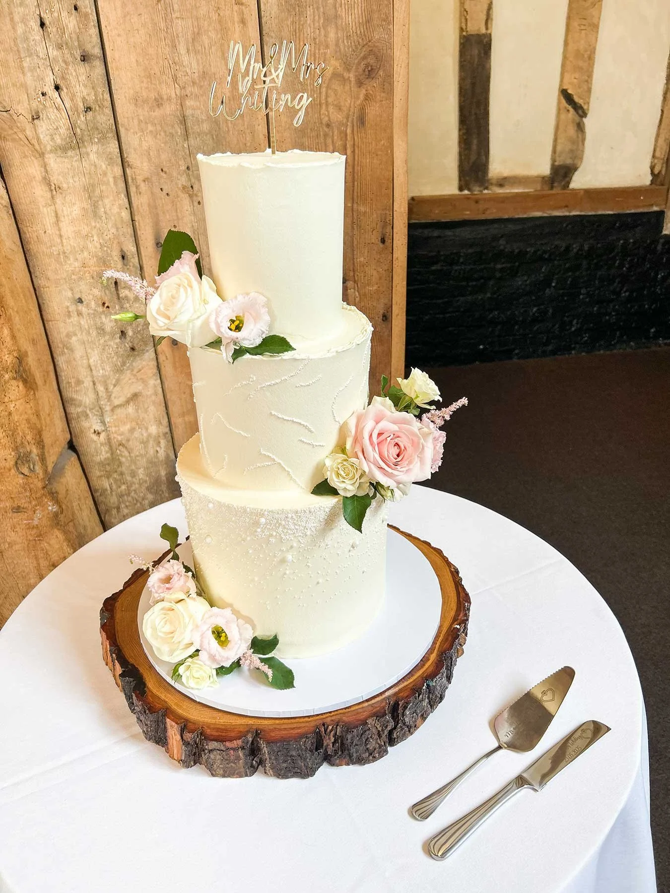 Four-tier wedding cake with white icing, decorated with pink and white roses and greenery, topped with 'Mr & Mrs Whiting' cake topper, on a wooden slab base, placed on a white table with cake knife and server.