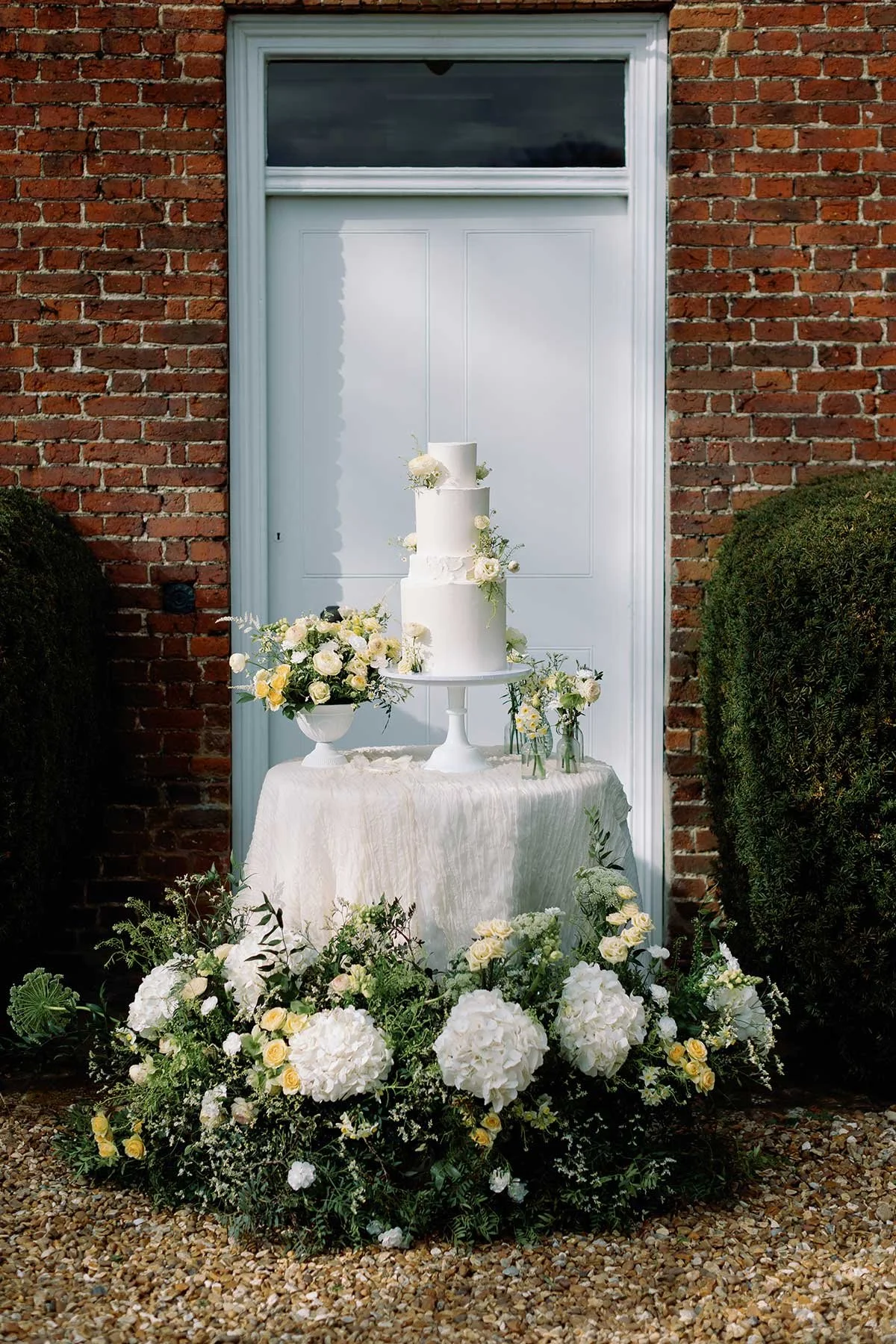 A white wedding cake with three tiers, decorated with white flowers, on a white pedestal table. Surrounding the table are white hydrangeas and other white and yellow flowers, all set against a brick wall and a white door.