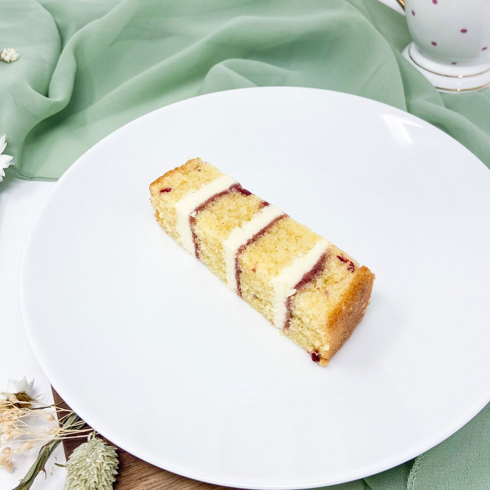 A slice of layered cake with cream and jam filling on a white plate, with a green cloth and flower decorations in the background.
