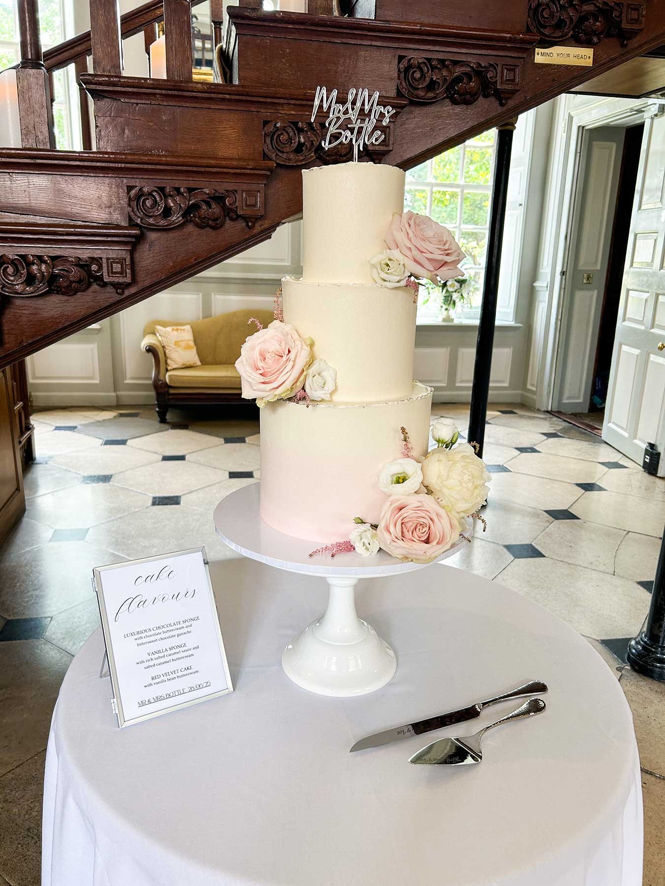 A three-tiered wedding cake decorated with pink and white roses and peonies, topped with a "Mrs & Mrs Bottle" cake topper, displayed on a white cake stand at a wedding reception.