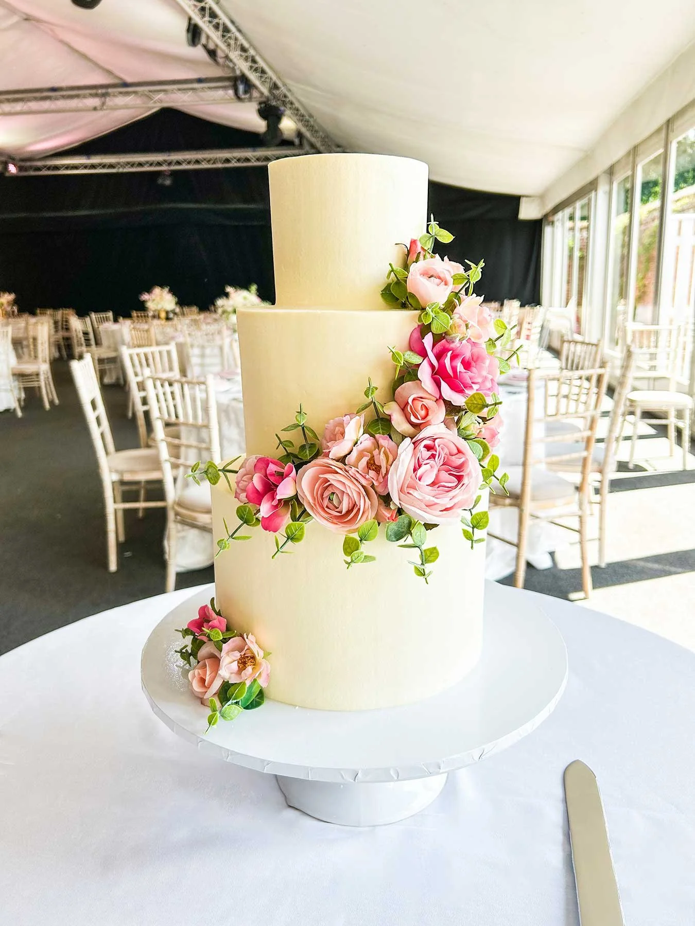 Two-tier wedding cake with white fondant, decorated with pink and blush roses and green leaves, placed on a white cake stand on a table draped with a white tablecloth.