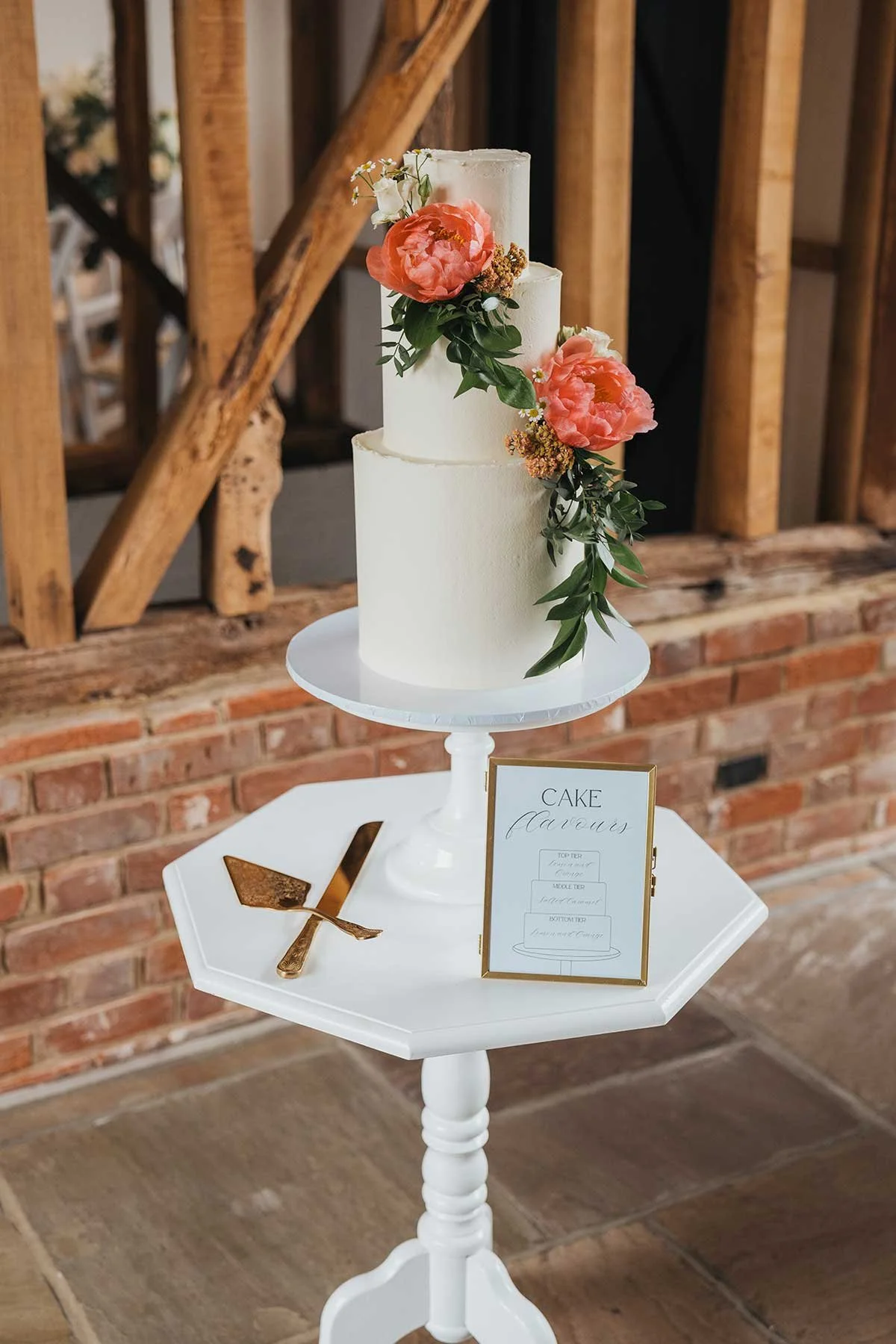 A three-tier white wedding cake decorated with pink and white flowers and greenery on a white cake stand. It is placed on a small white table with a cake slicing set and a framed sign.