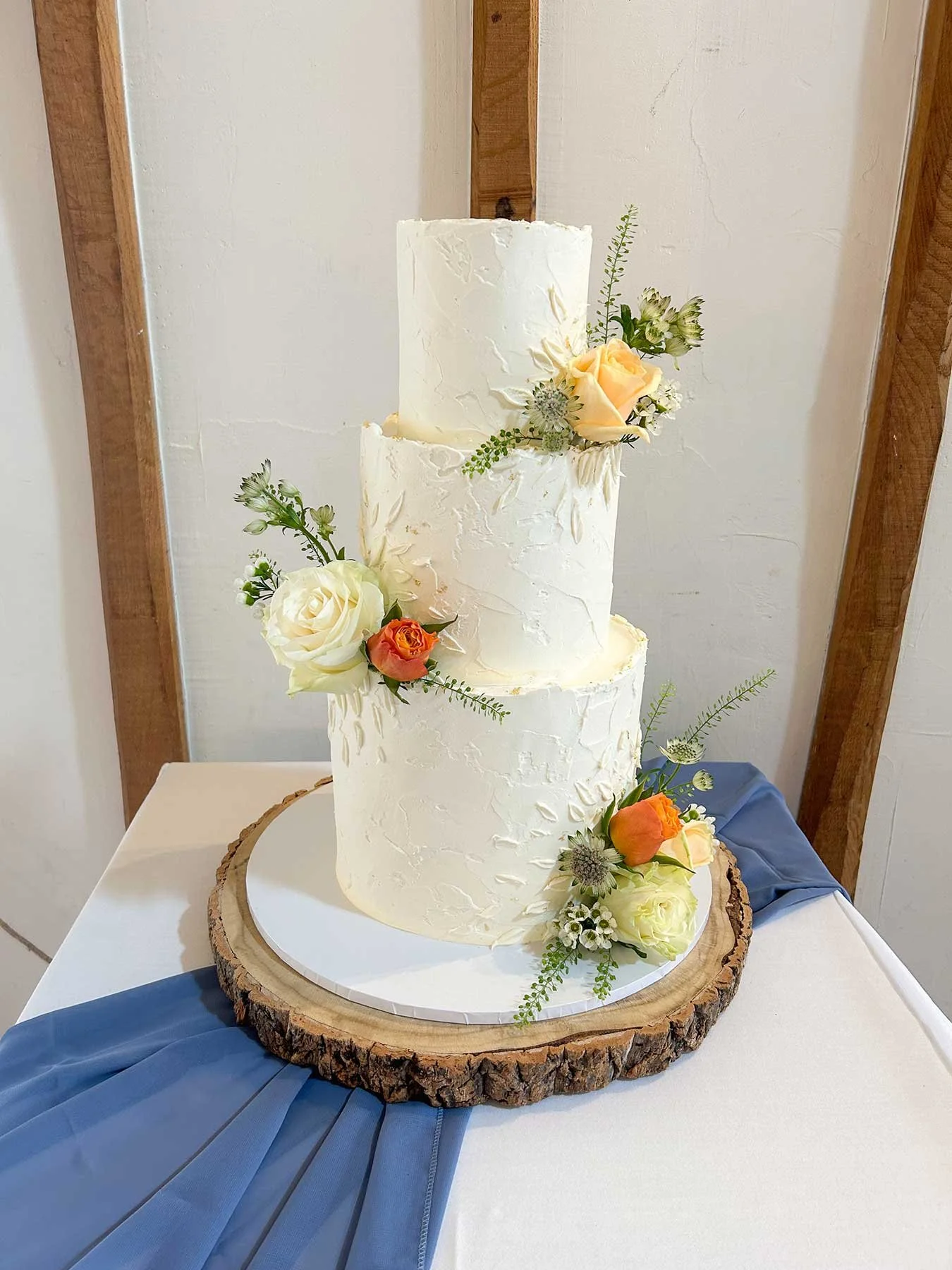 Three-tier white wedding cake with textured icing, decorated with cream, peach, and orange roses and greenery, placed on a wooden cake stand with blue drape underneath.