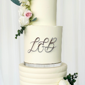 A white wedding cake with pink and white roses and green foliage, decorated with the initials 'LGB' in purple script.