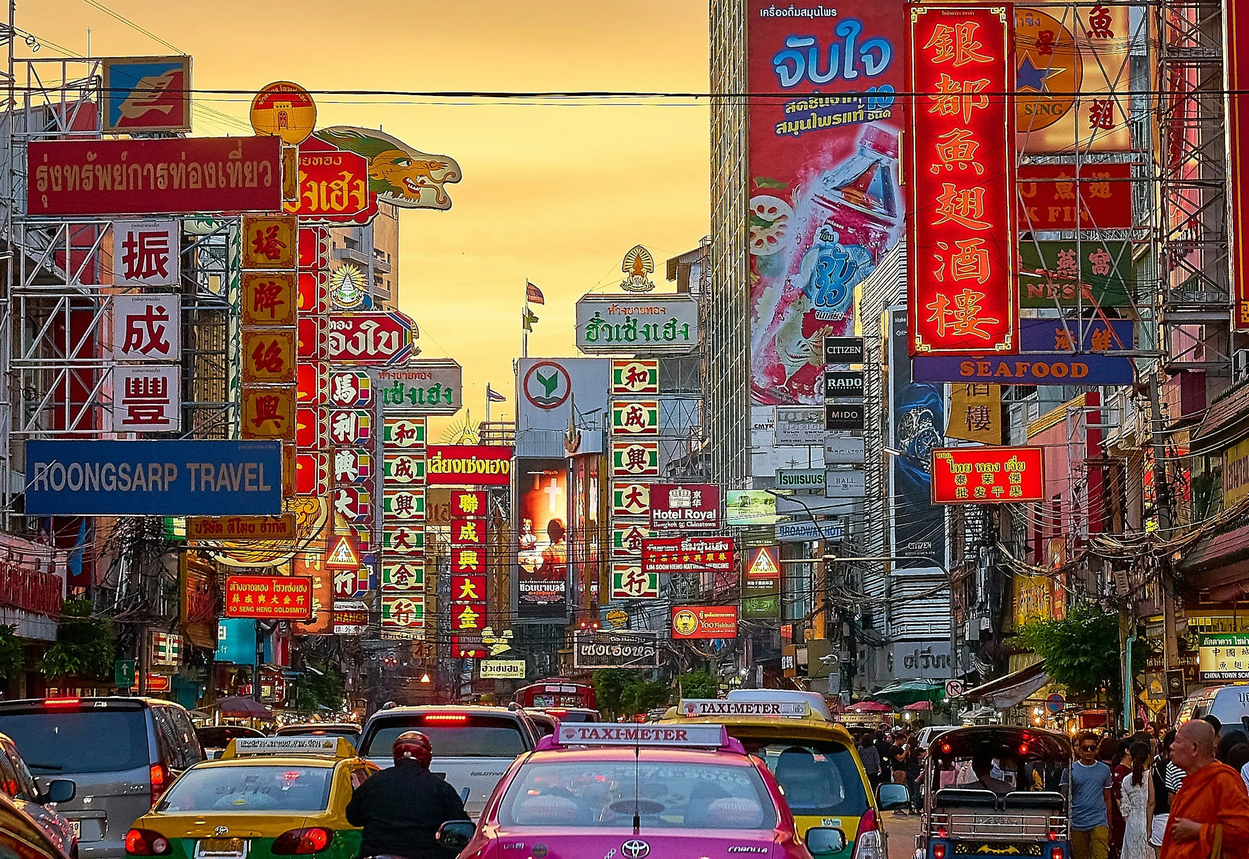 A busy street scene in an Asian city with numerous colorful neon signs in multiple languages, traffic, and pedestrians during sunset.