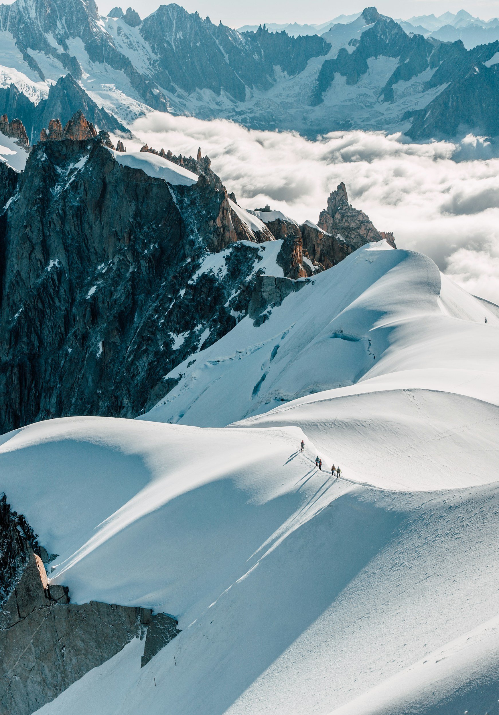 Four climbers ascending a snow-covered mountain ridge with rugged, rocky peaks and clouds in the background.