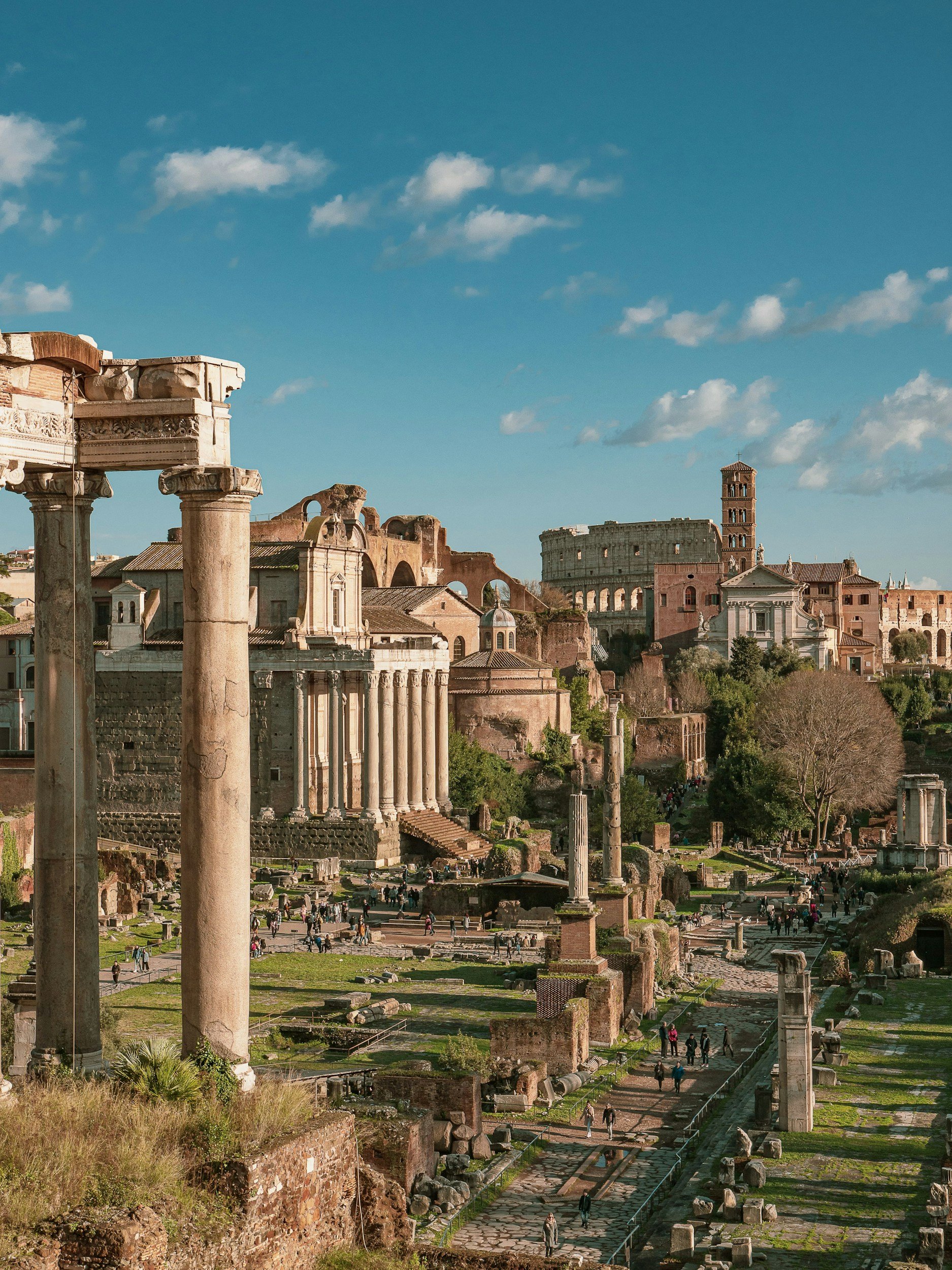 Ancient ruins of the Roman Forum with standing columns, arches, and historic buildings under a blue sky with clouds.