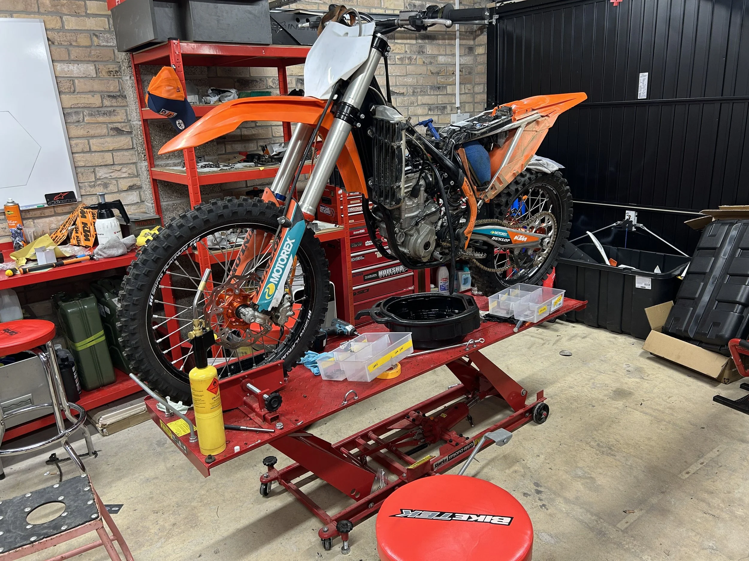 A motocross motorcycle on a red work table in a workshop, with parts and tools scattered around, some of the motorcycle's body panels removed.