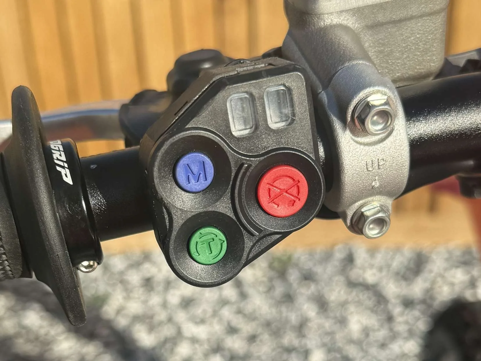 Close-up of motorcycle handlebar control buttons: blue 'M', red 'X', and green 'T' buttons, with a textured black grip and metal seat clamp.