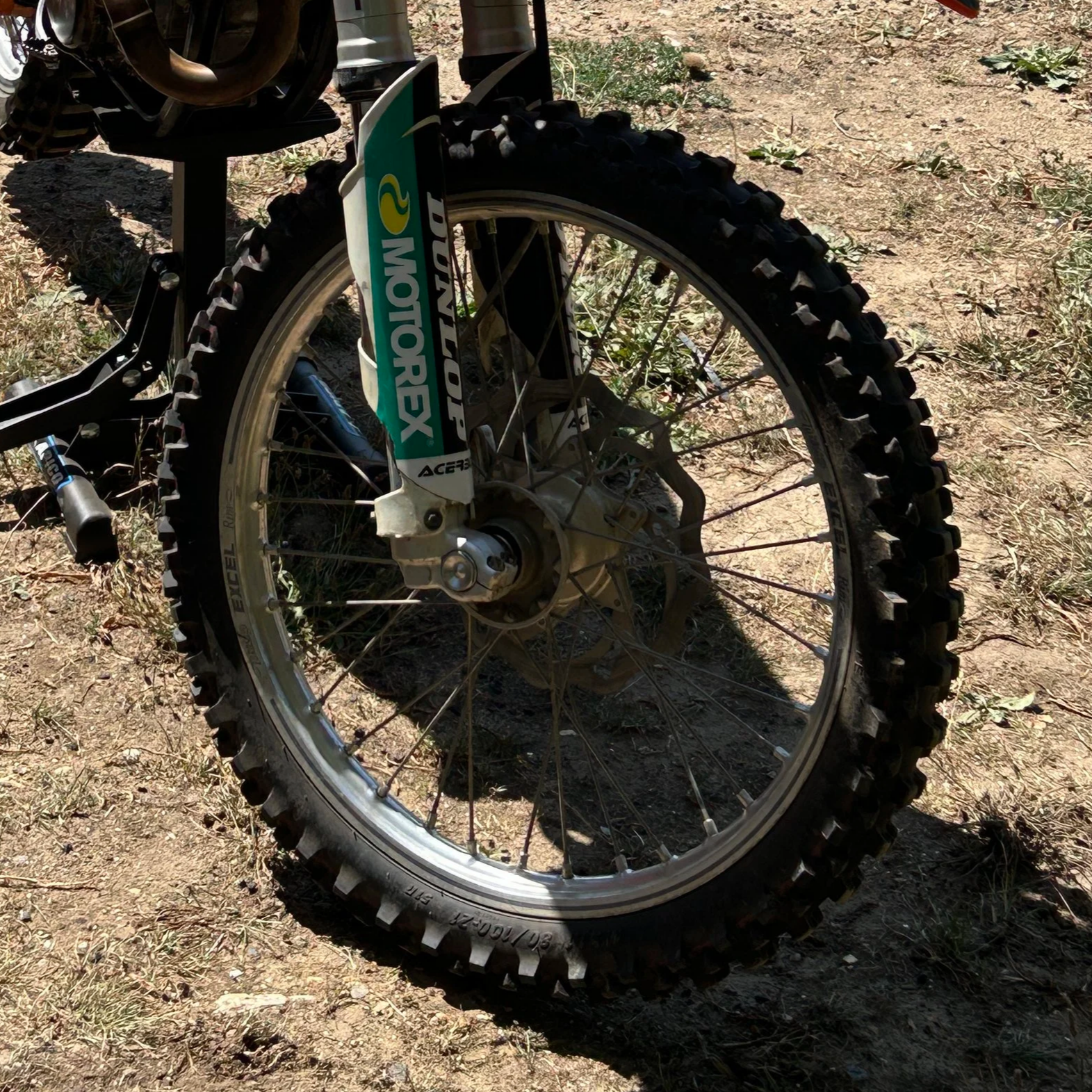 Close-up of a dirt bike front wheel on a dirt trail, showing knobby tire, spokes, and part of the suspension fork with a Motorex decal.