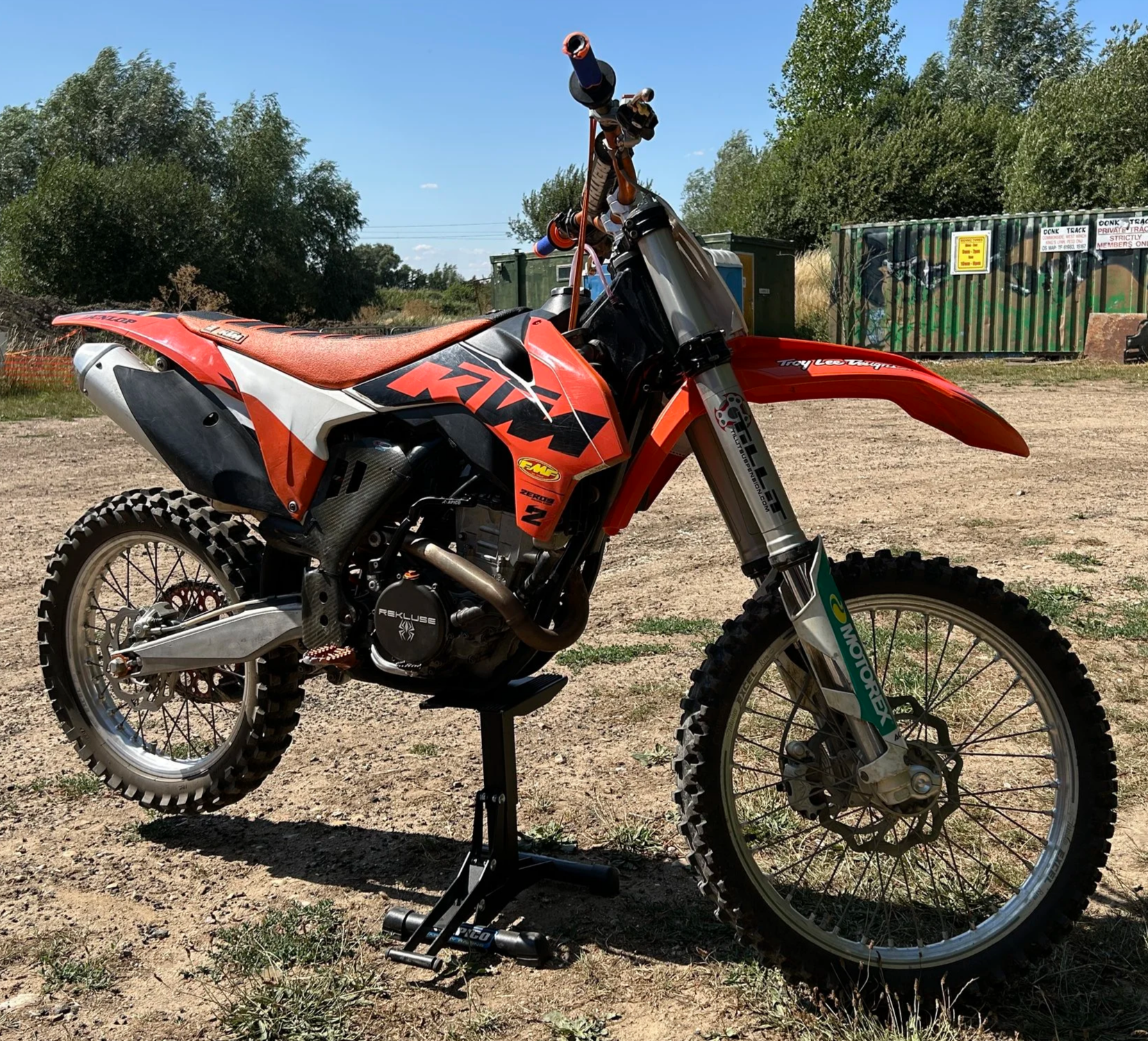 Orange and black motocross bike on stand outdoors on dirt ground with trees and shipping containers in background.