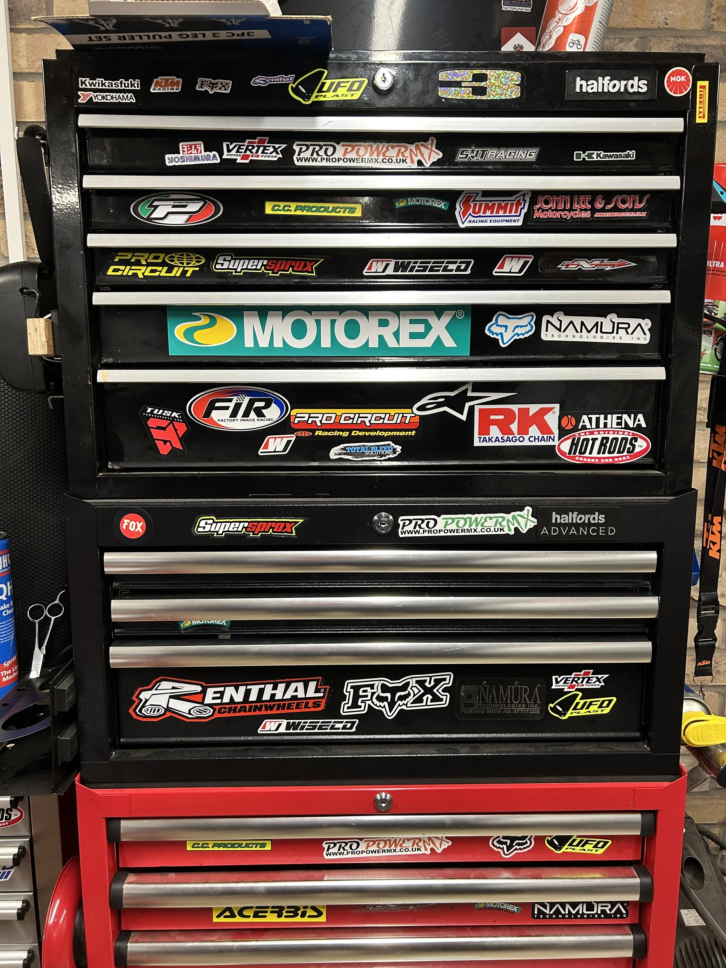 A black and red tool chest with multiple drawers, covered with various motorcycle and racing brand stickers, including Motorex, Fox, Kawasaki, and Pirelli.