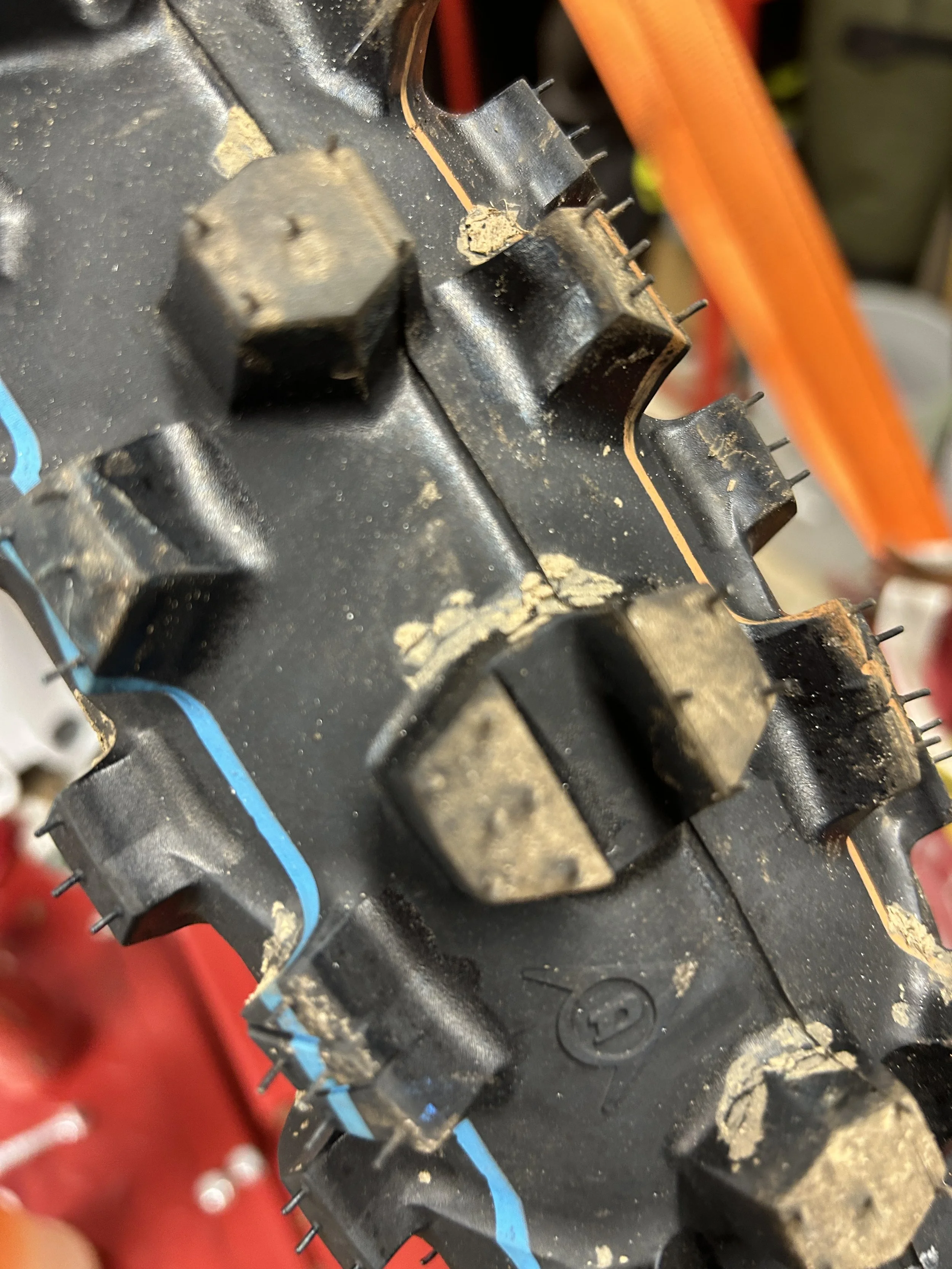 Close-up of a black rubber tread of a tire with dirt and mud on the surface. The tire has large, rugged lugs and colored lines on the sidewall.
