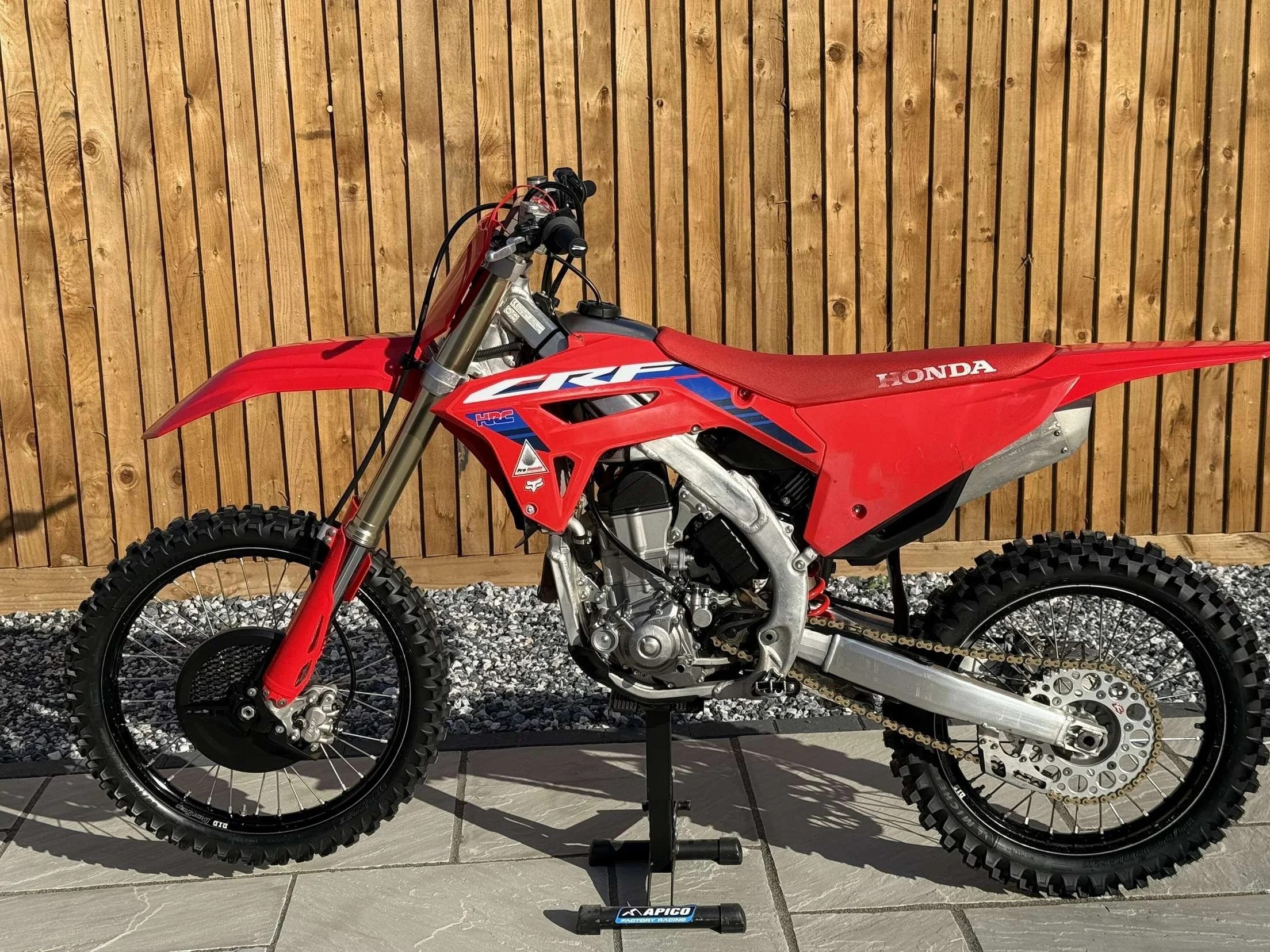 Red Honda off-road motorcycle with knobby tires, displayed on a black stand in front of a wooden fence.