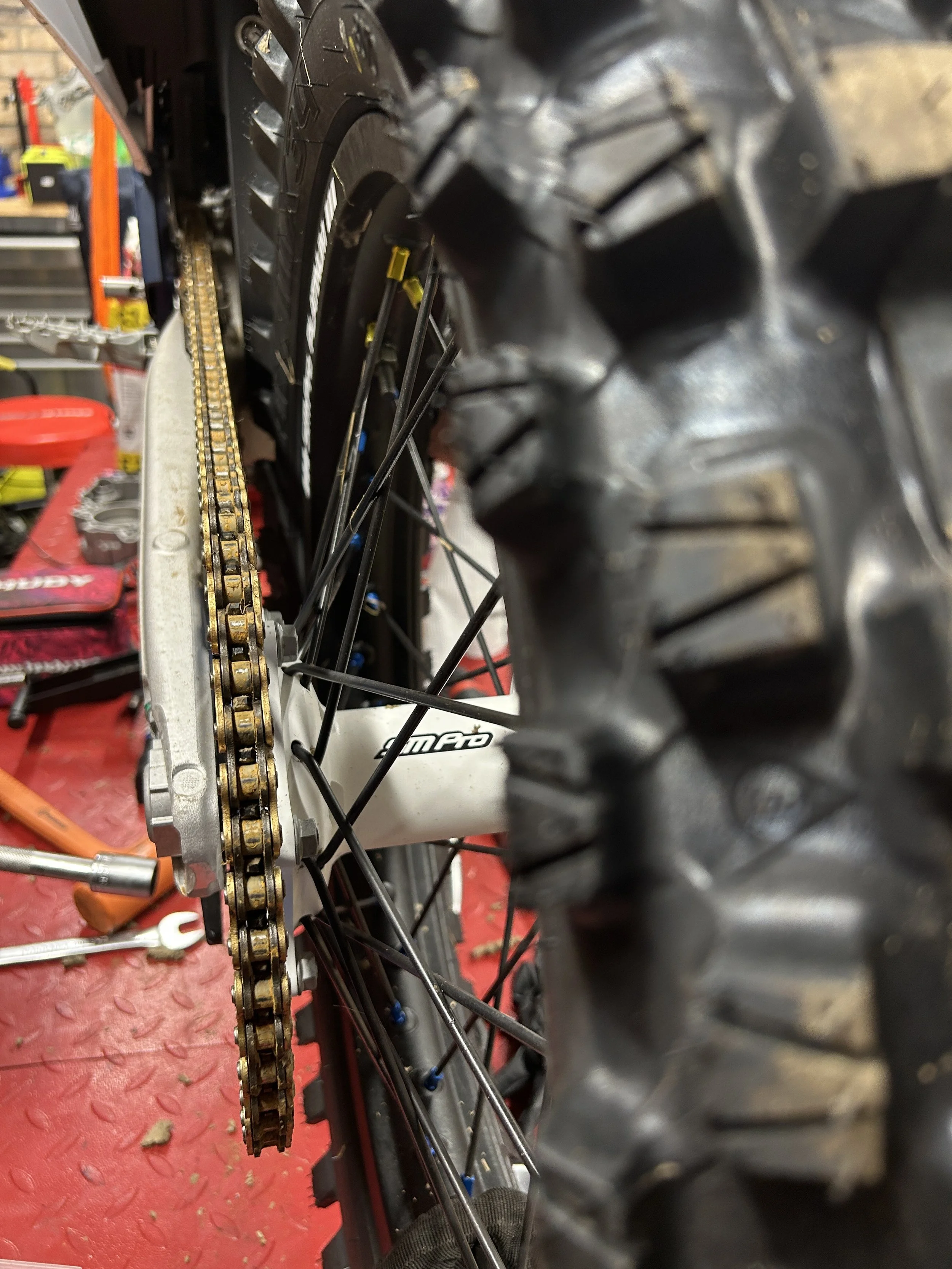 Close-up of a bicycle's rear wheel, chain, and tire in a workshop setting with tools and equipment in the background.