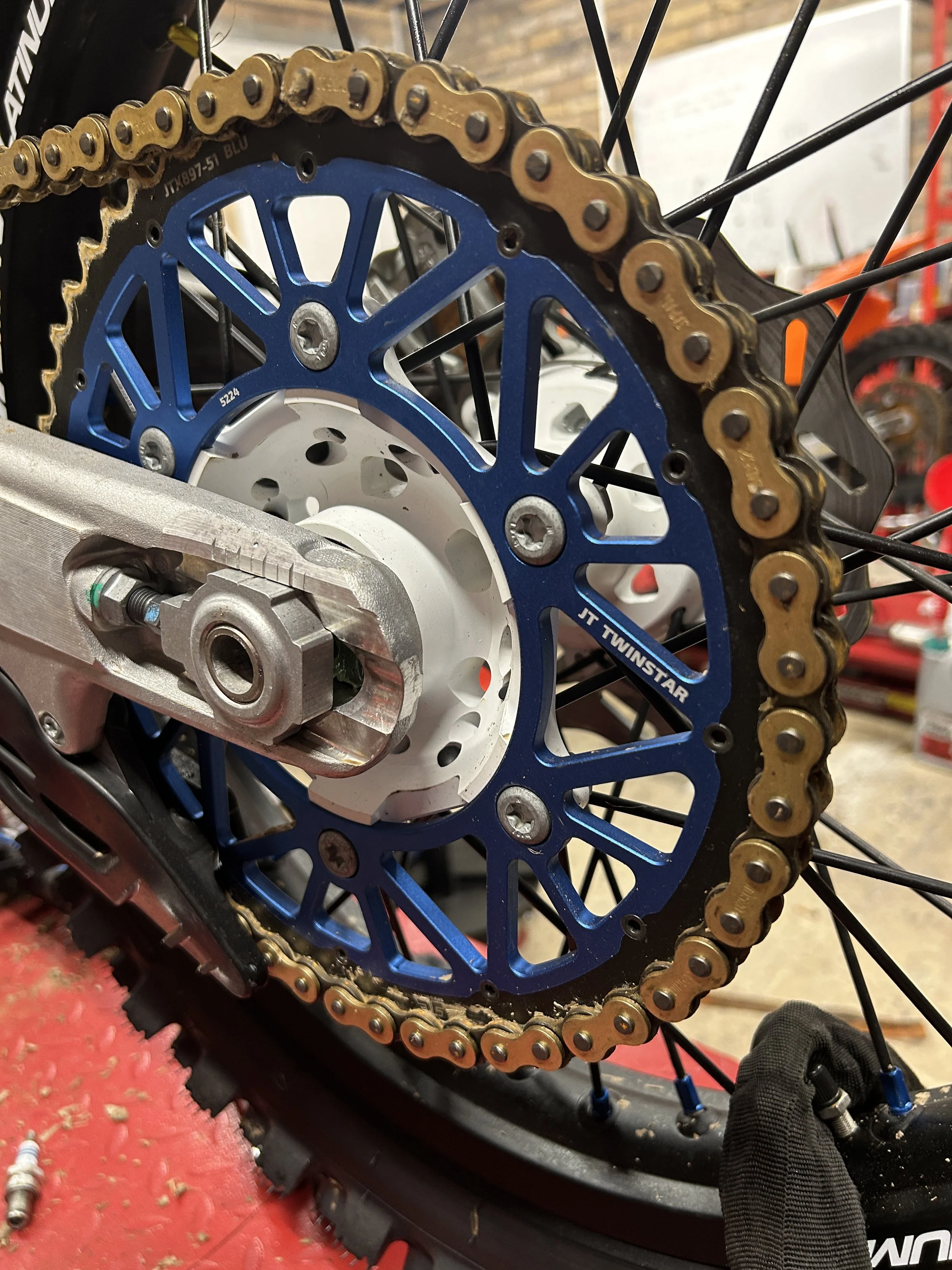 Close-up of a bicycle's rear sprocket and chain, with black spokes, and part of the wheel and hub visible, on a red workbench.