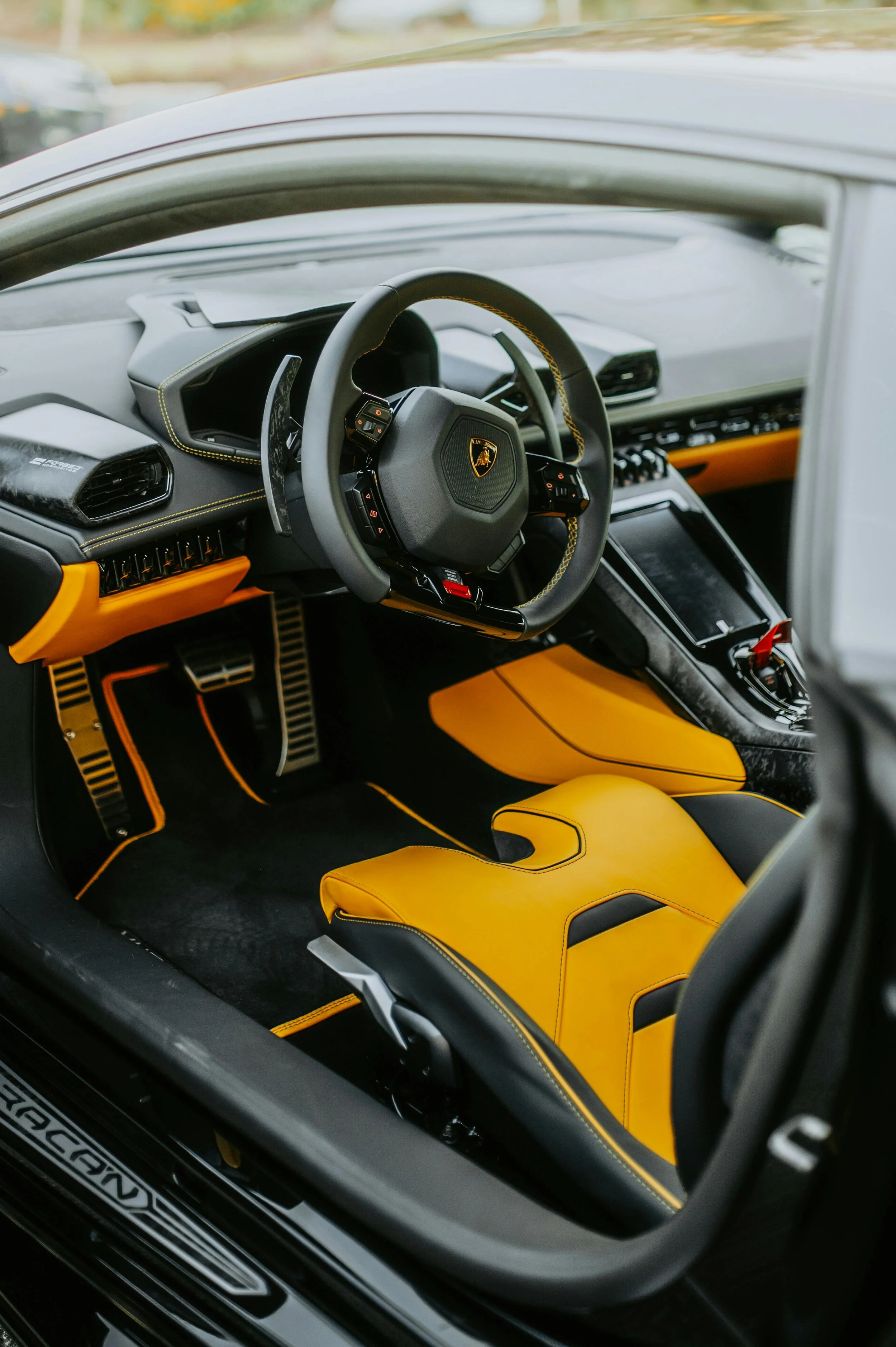 Interior of a luxury sports car with black and yellow accents, including a yellow seat, modern dashboard, and steering wheel with a Lamborghini emblem.
