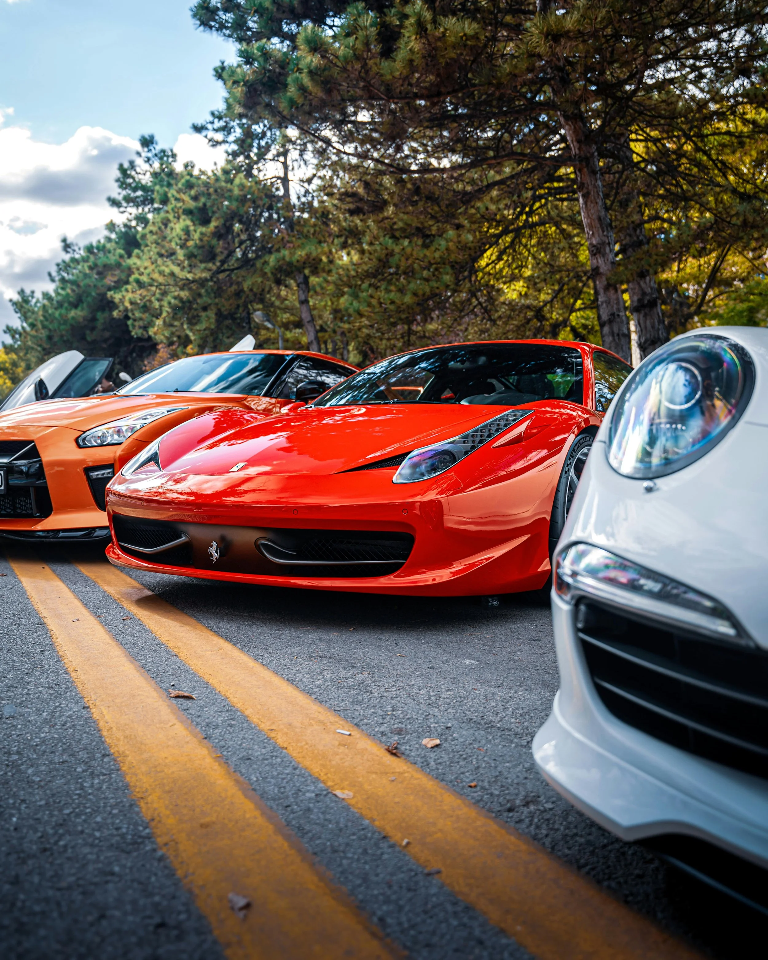 Row of luxury sports cars parked on a street with trees overhead, featuring a red Ferrari in the center, flanked by a white and an orange car.