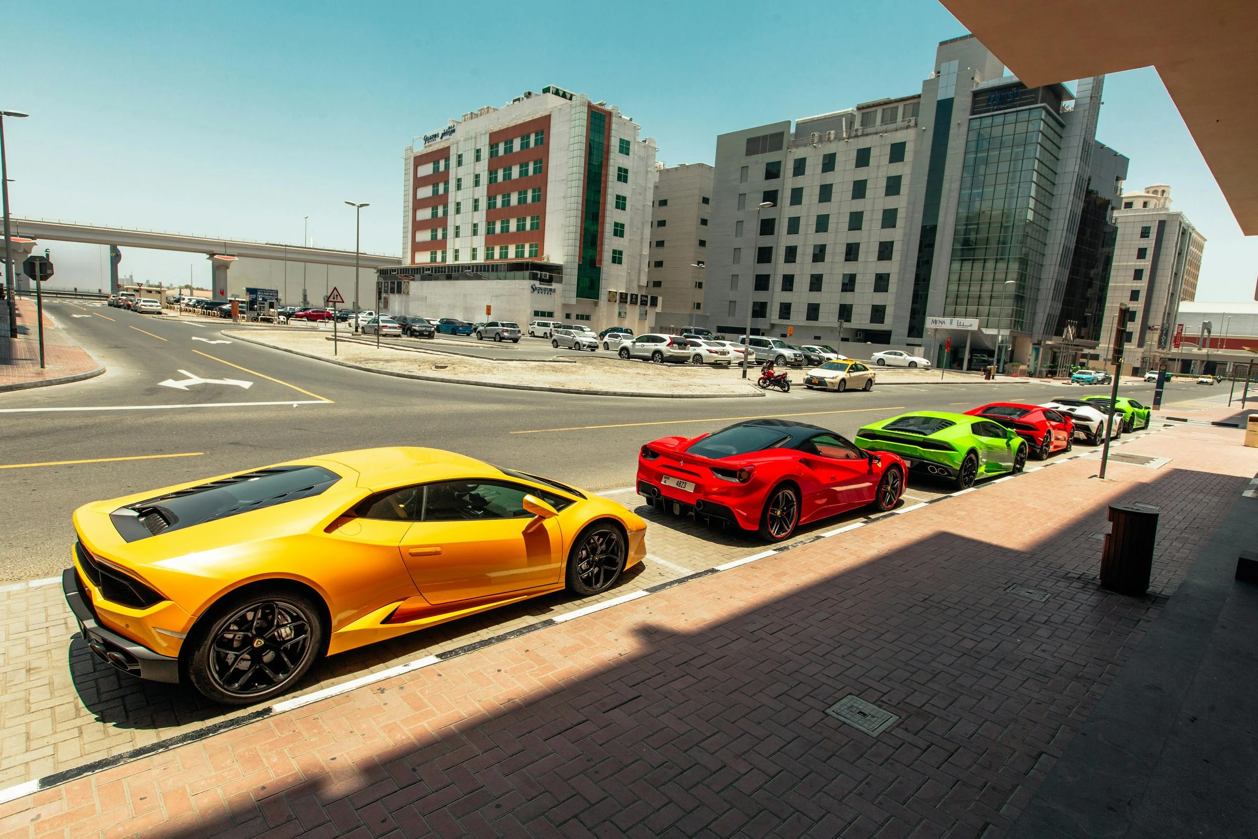 Four luxury sports cars parked along a city street on a sunny day, including a yellow Lamborghini, a red Ferrari, a green Lamborghini, and a red car, with modern buildings in the background.