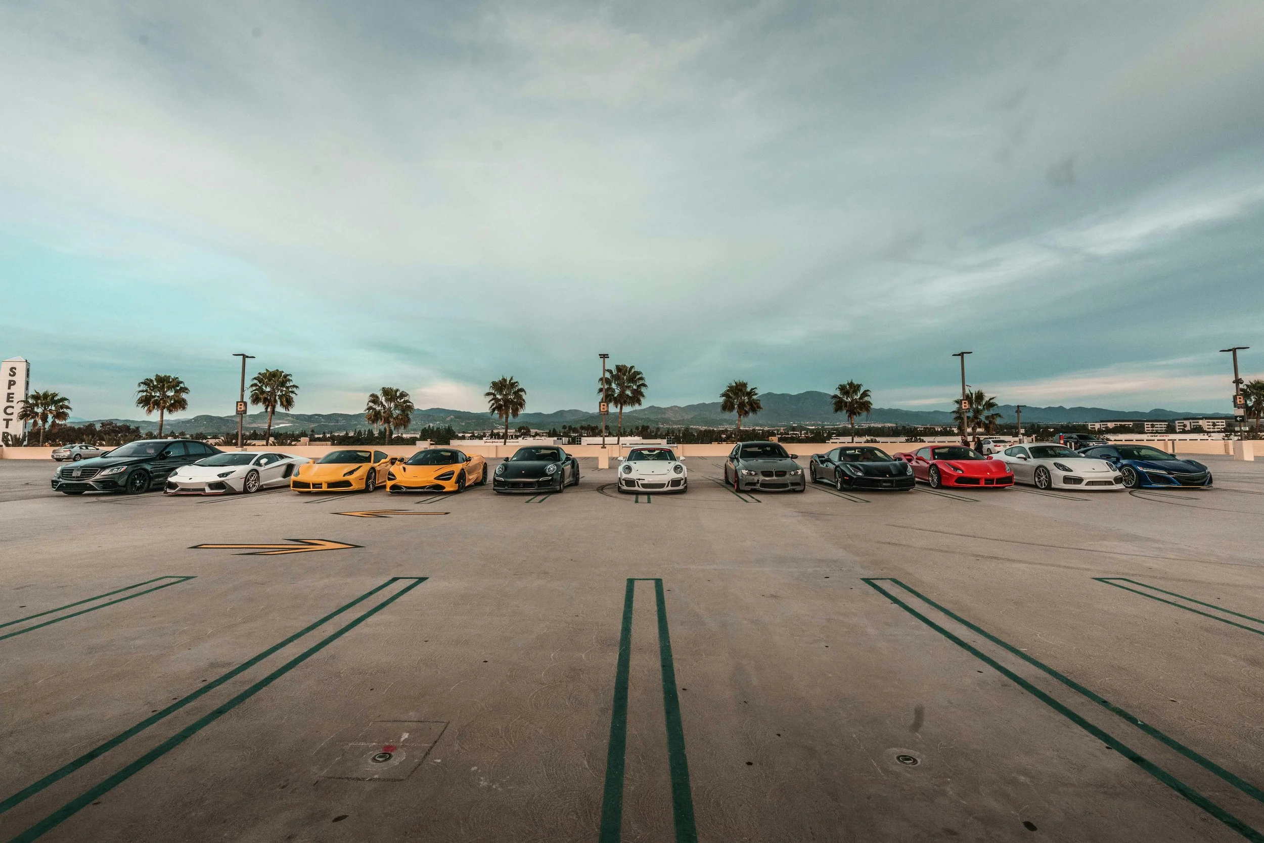 A row of various sports cars parked in a parking lot with palm trees and mountain range in the background.