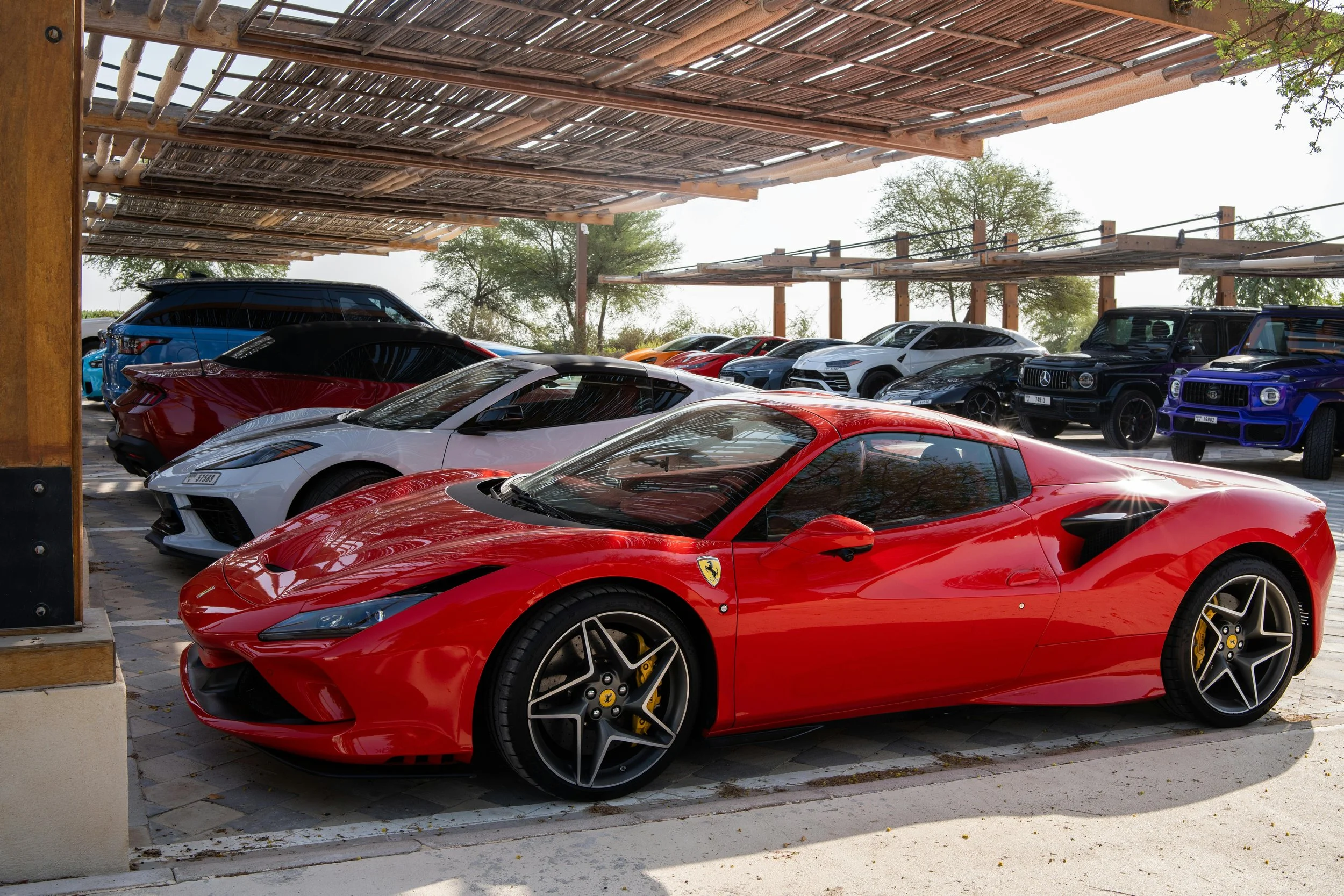 A red Ferrari sports car parked among other luxury vehicles in an outdoor lot with a shaded wooden structure and trees in the background.