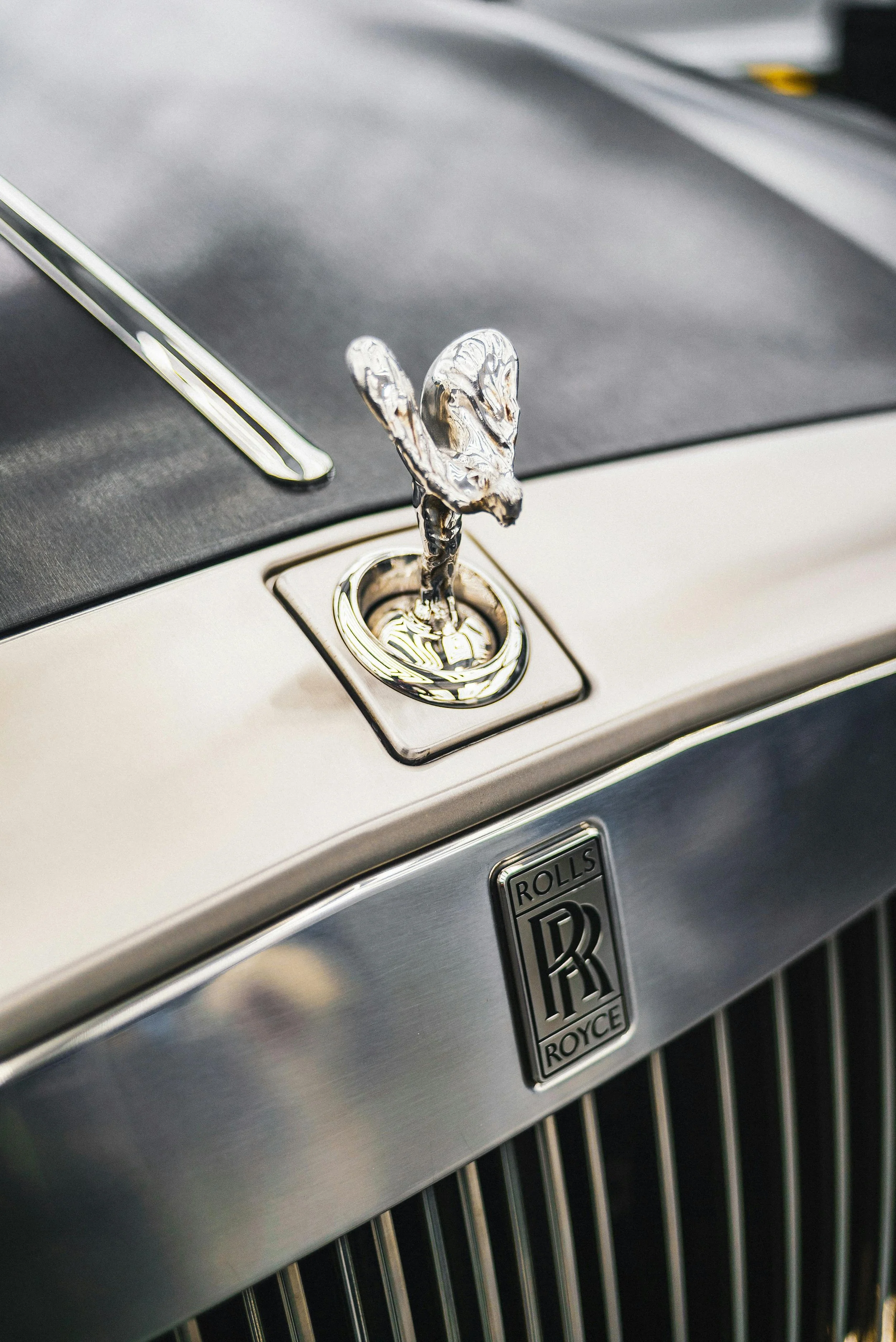 Close-up of a vintage Rolls-Royce car hood ornament, featuring a silver Spirit of Ecstasy figure mounted on the grille with the Rolls-Royce emblem below.