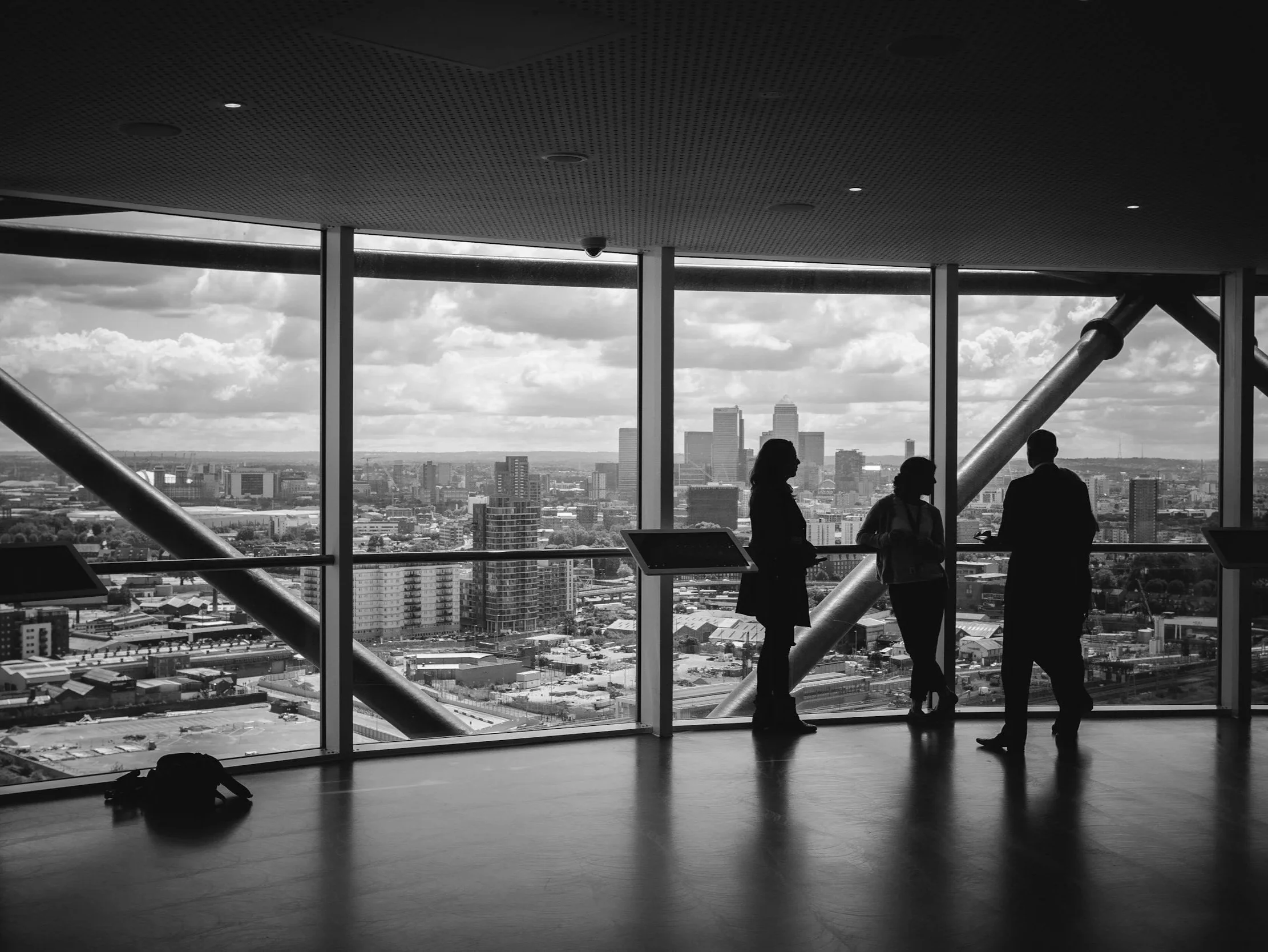 Silhouettes of three people standing and talking inside a modern high-rise building with large floor-to-ceiling windows, overlooking a city skyline with tall buildings and partly cloudy sky.