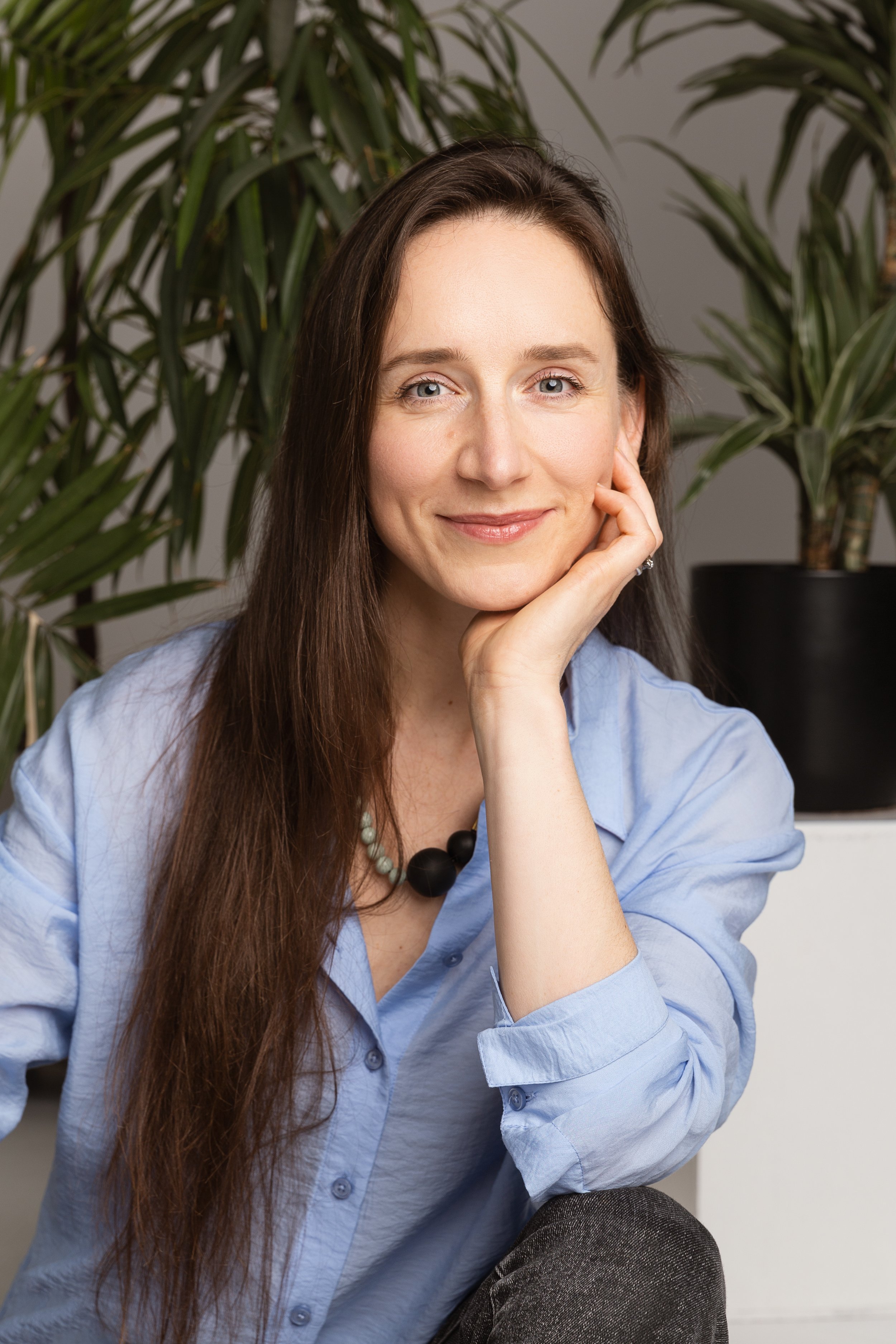 Psichologė Urtė Navickienė, dėvinti šviesiai melsvus marškinius, sėdi prieš žalius augalus ir maloniai šypsosi.