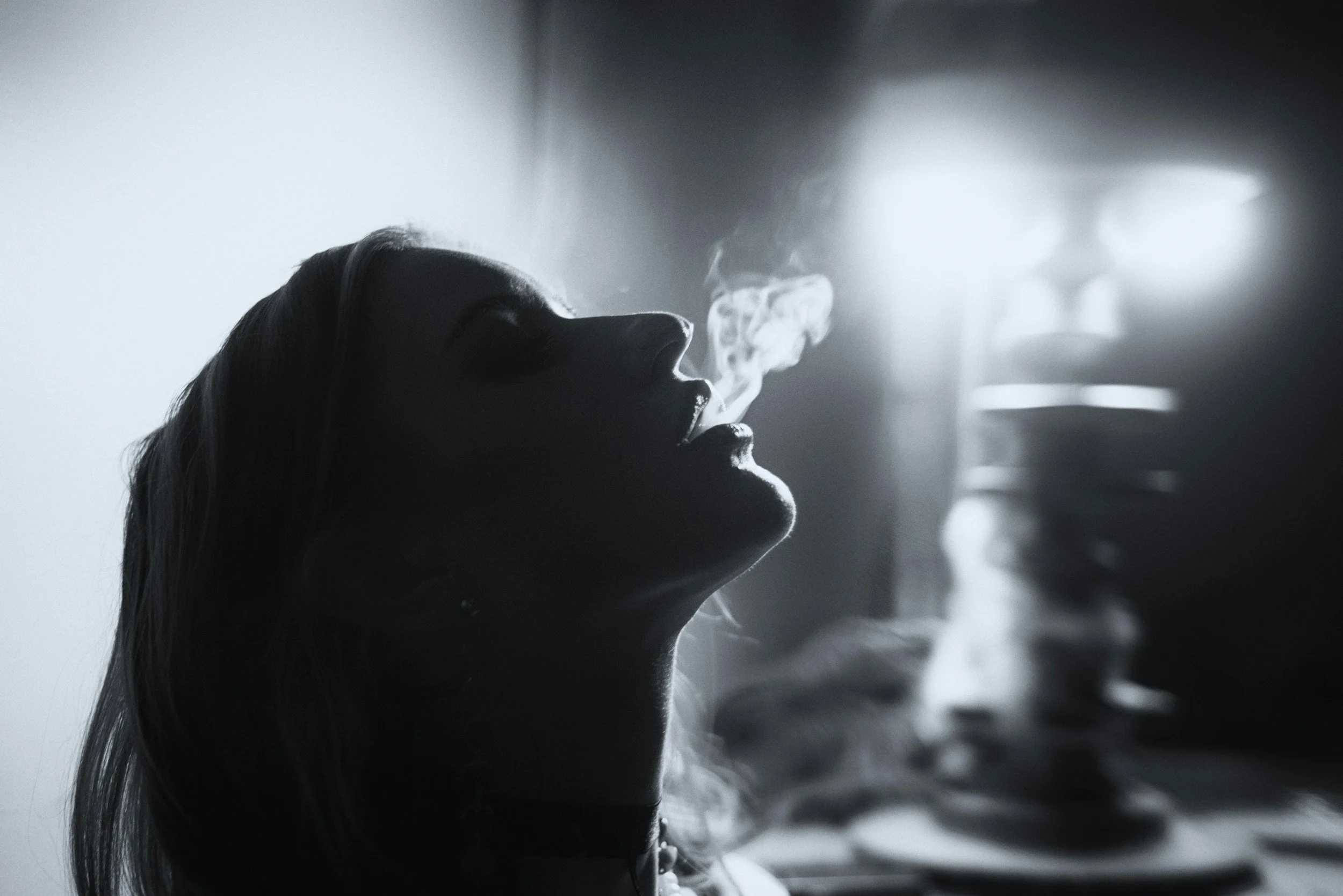 A black and white photo of a woman with long hair, breathing out smoke or vapor in a dimly lit room with a soft light behind her.