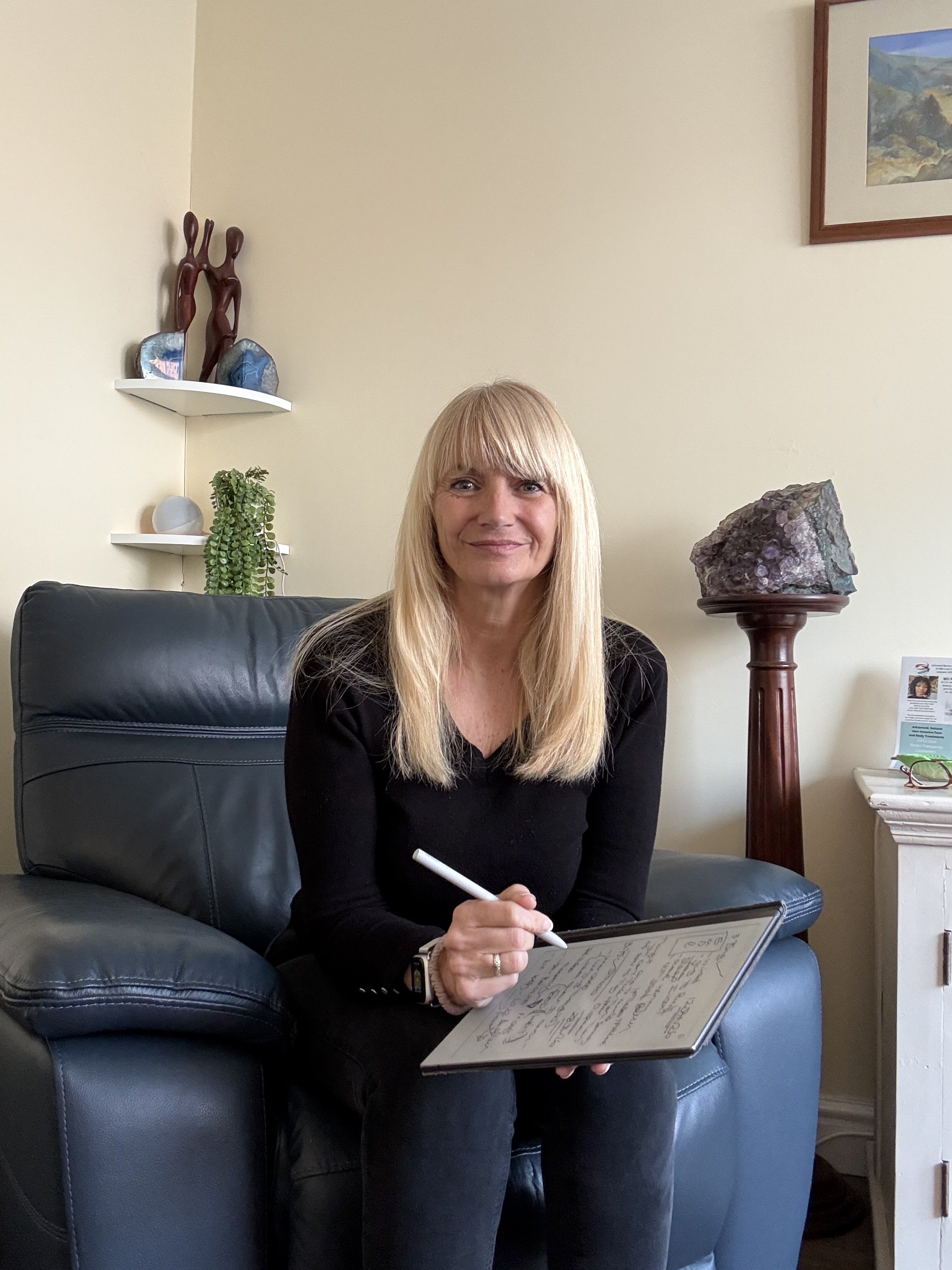 A woman with long blonde hair sitting on a black leather couch, holding a stylus and writing on a digital tablet, in a room with light-colored walls, decorative shelves, and a large mineral on a wooden stand.