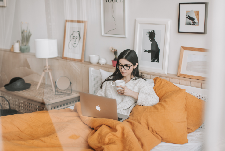 A young woman with glasses sitting on a bed with orange bedding, working on a silver MacBook laptop and holding a white mug in her hand.