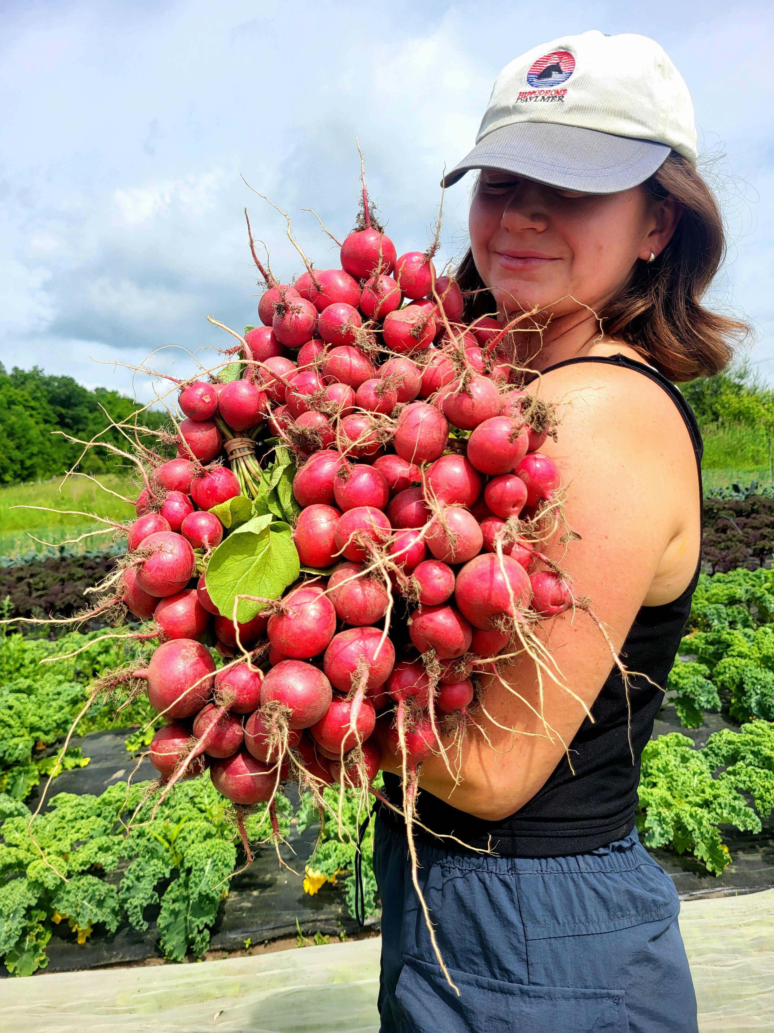 Woman in a hat holding a large bunch of radishes in a farm field.