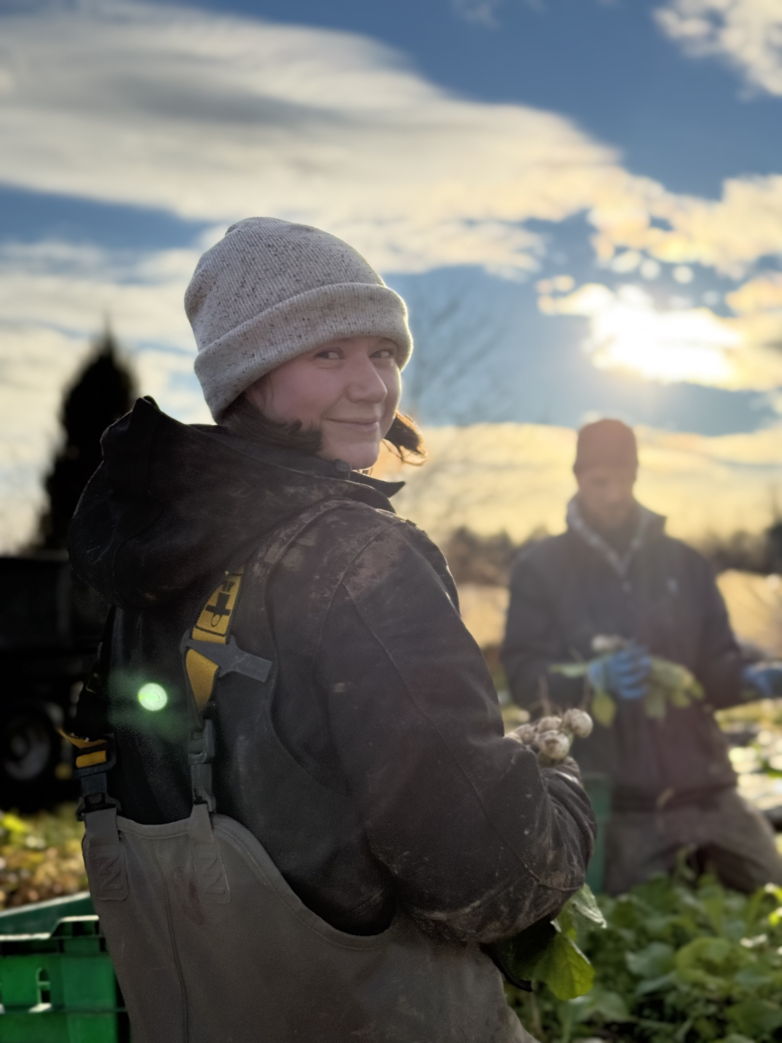 A woman wearing a gray knit hat and black jacket holding fresh vegetables outdoors during sunset, with a smiling face, and another person in the background harvesting vegetables.