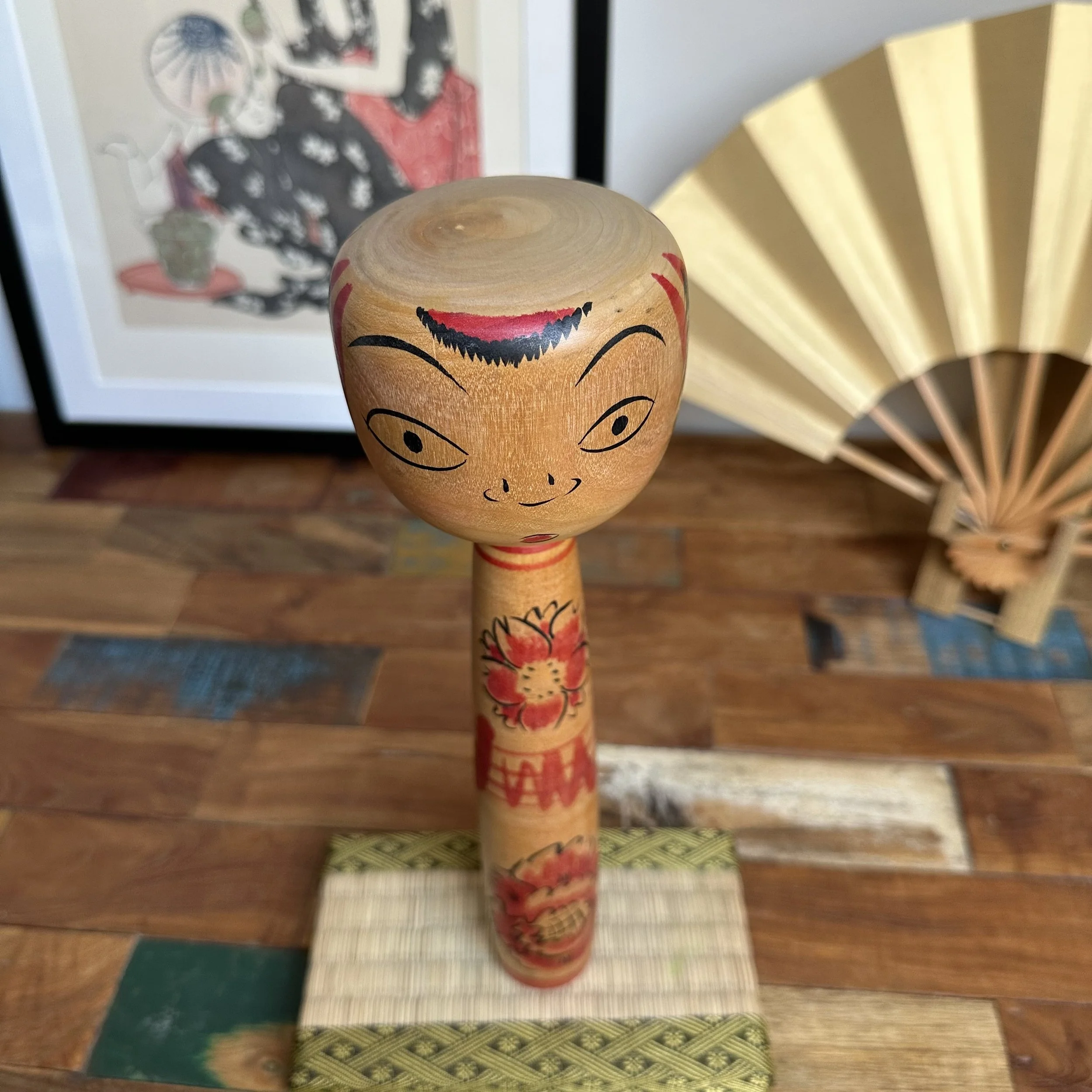 Seya Jūji kokeshi doll with a large, expressive face, black eyes, red forehead decoration, and body painted with red cherry blossoms, on a woven mat.