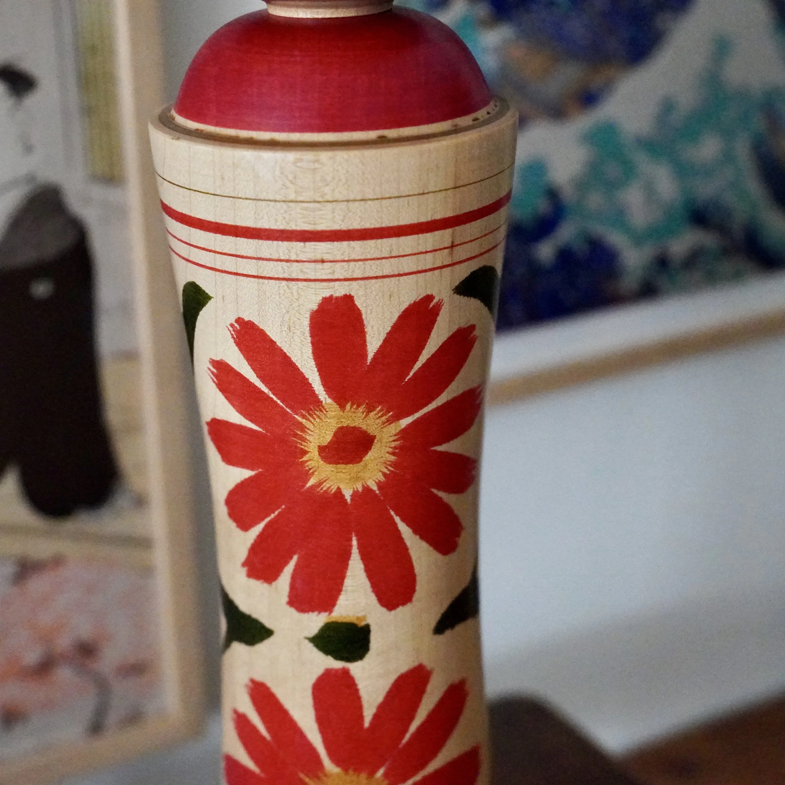 Close-up of a Ginzan kokeshi by Izu Mamoru, with a red domed top, pale wooden body featuring red stripes, and large red chrysanthemums with yellow centers.