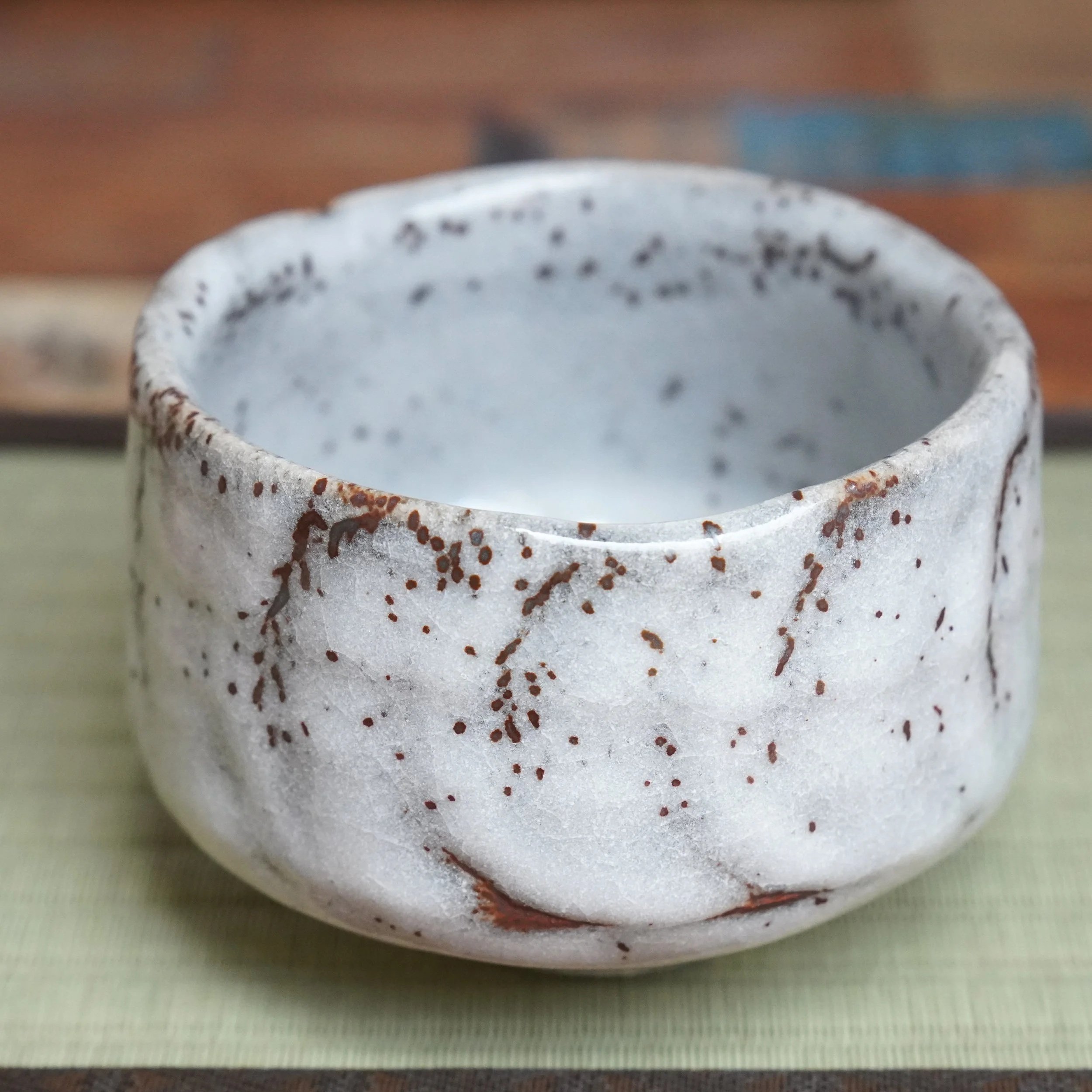 Katō Shōzan's Oni-Shino chawan, a white ceramic bowl with irregular shape, crackled glaze, and rust brown specks and drips, on a tatami mat.