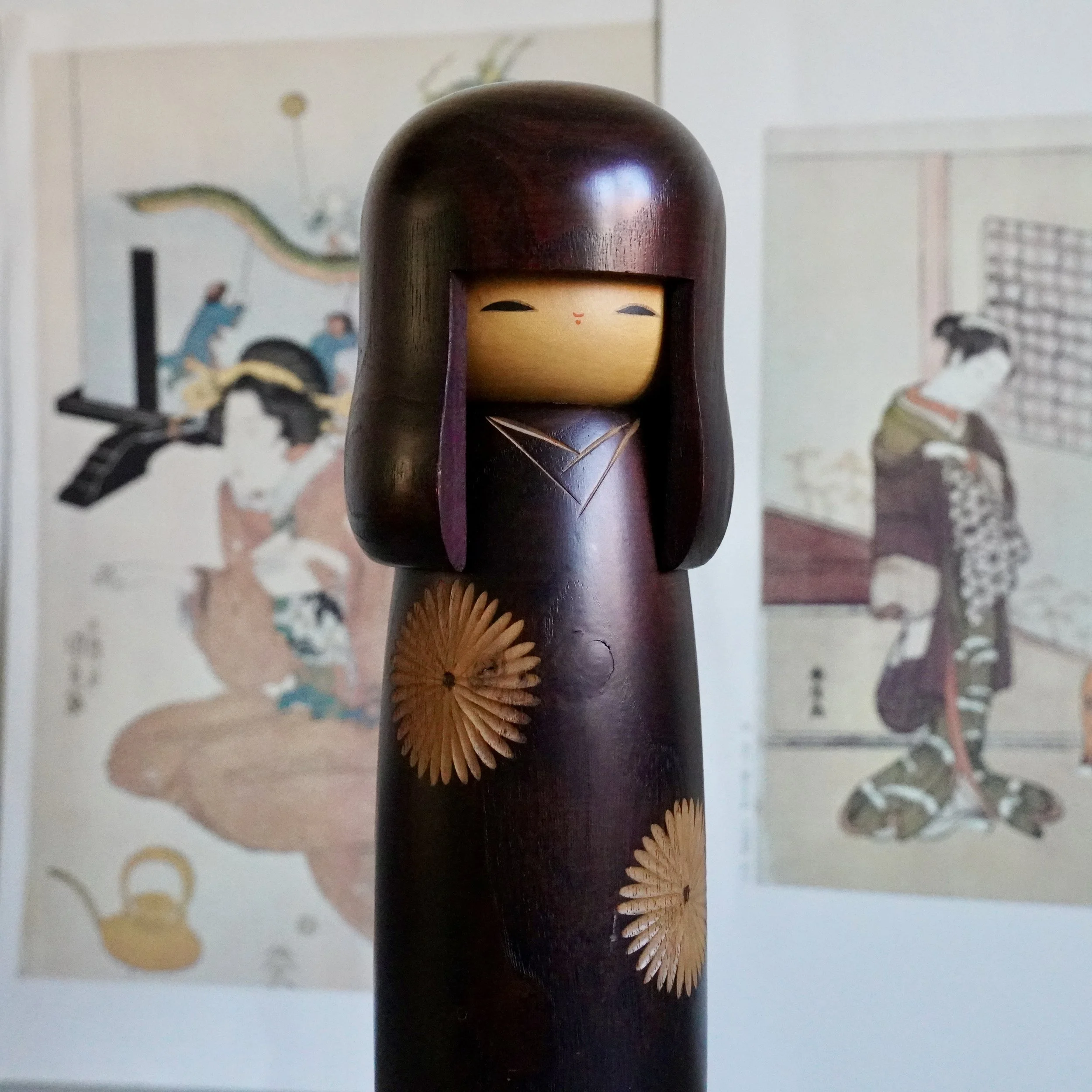 Sosaku kokeshi close angle view, aubergine stained wood, downcast painted face, chrysanthemum inlay detail