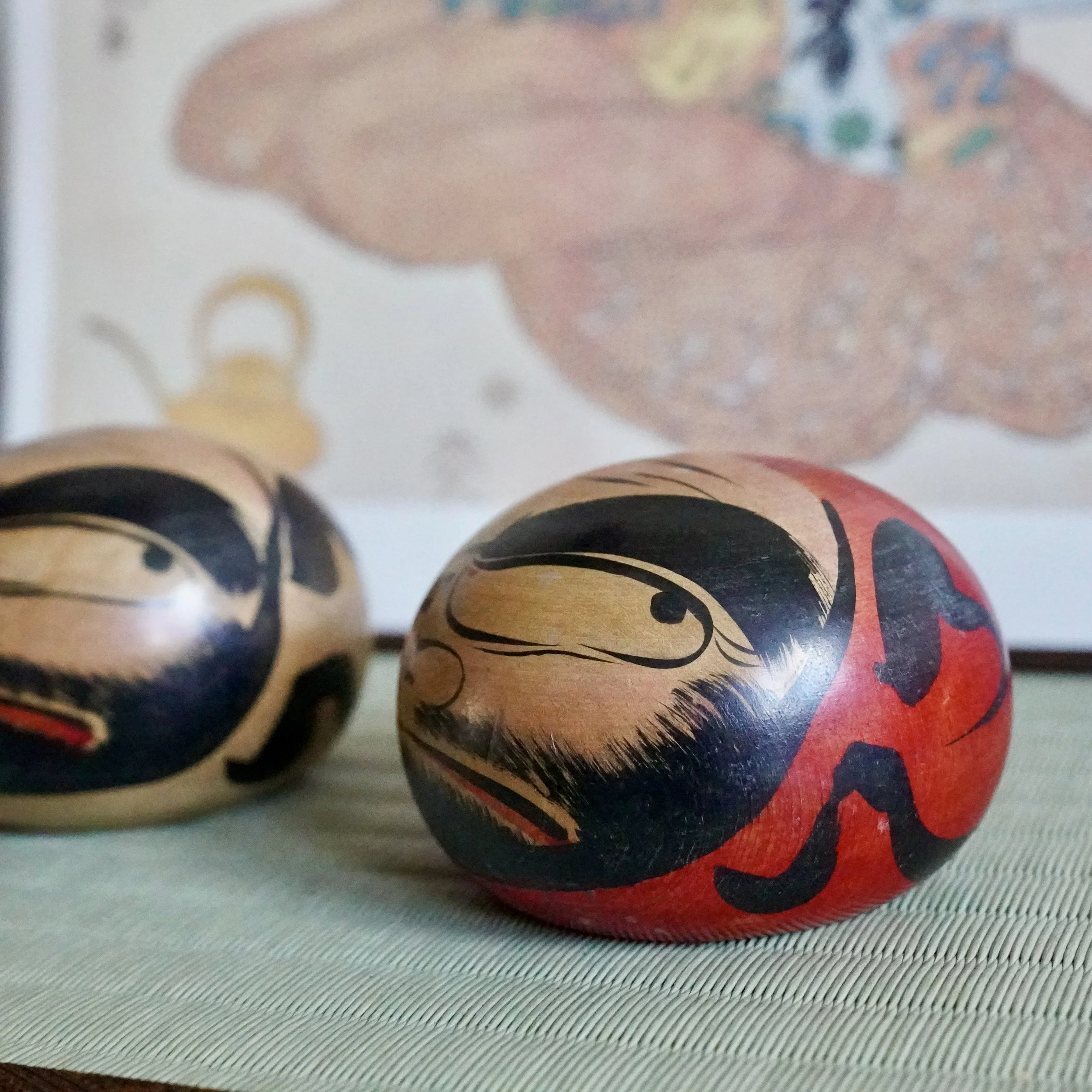 Close-up of two Tsugaru daruma by Satō Zenji; one red and black, the other natural wood, both with hand-painted scowling faces, resting on a woven mat.