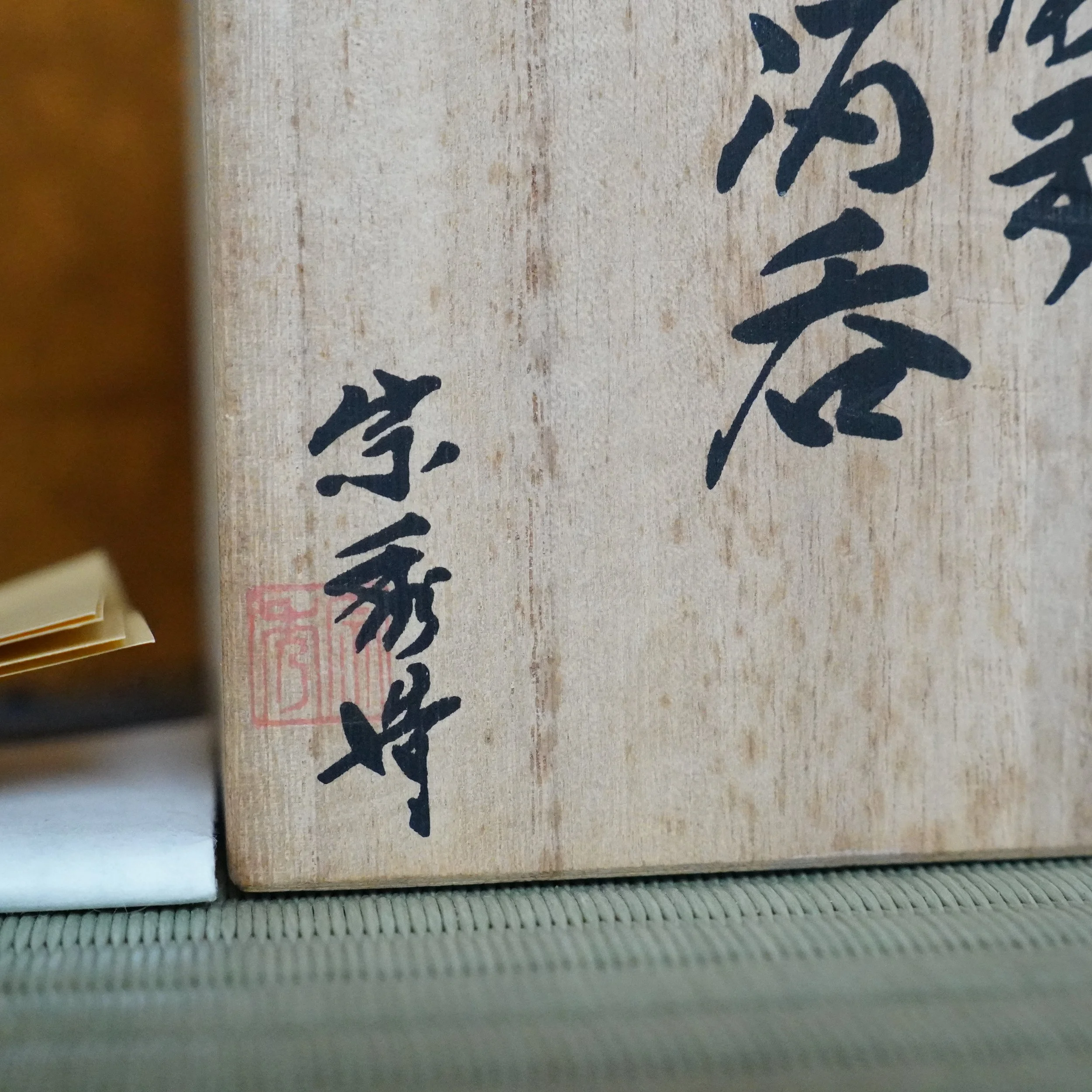 Wooden tomobako box for a Bizen-yaki guinomi, signed Yamamoto Sōshū in black calligraphy with a red seal, resting on a woven mat.