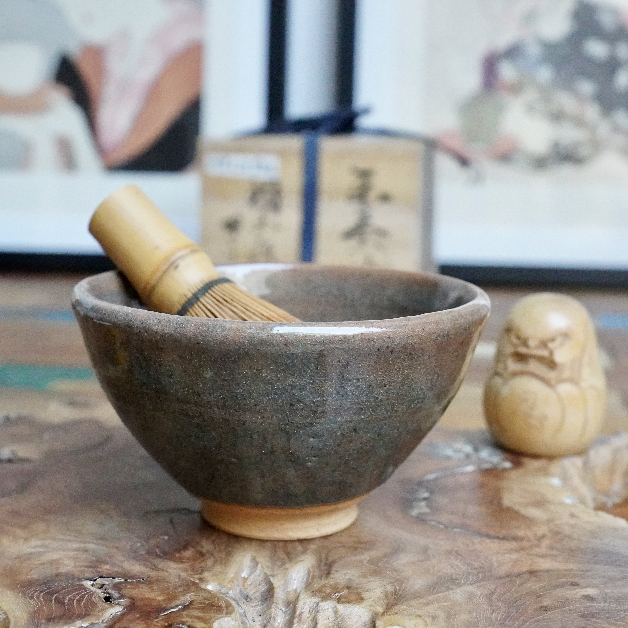 Mizushima Jirō's Kagurao-yaki blue chawan, a textured stoneware bowl with a bamboo tea whisk, on a wooden surface. A figurine and tomobako are in the blurred background.