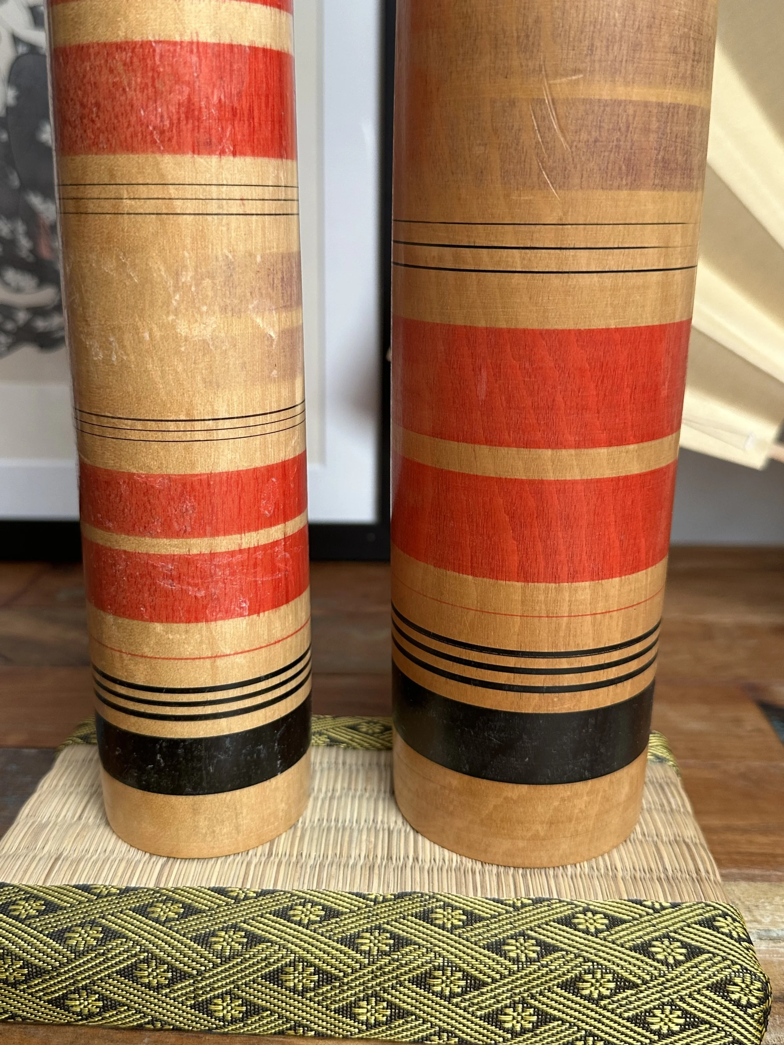 Two wooden Tsuchiyu kokeshi with horizontal bands of red, black, and natural wood, side by side on a woven mat with a patterned border.