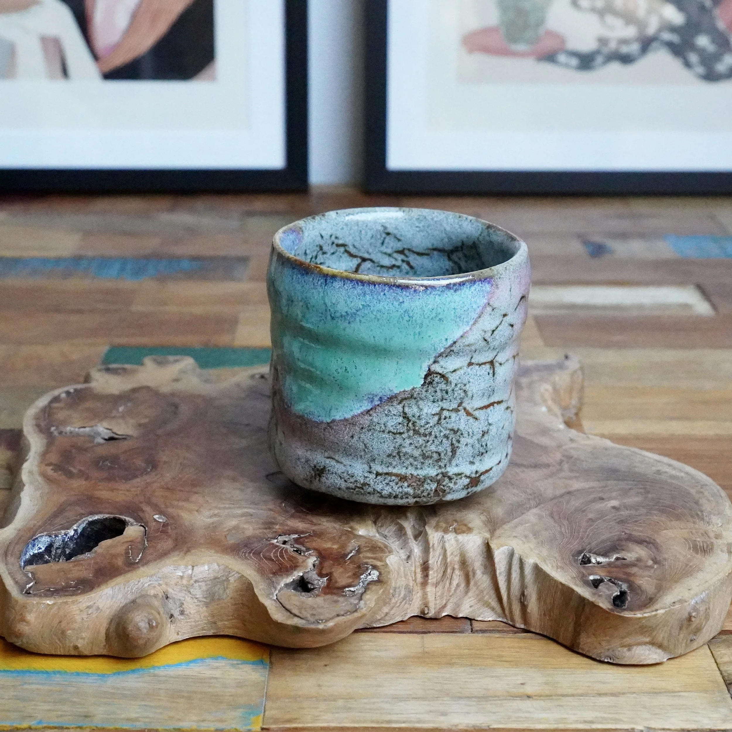 Tsutsu chawan with layered turquoise and speckled grey glaze, featuring warm brown crackle lines, resting on an organic wood slab.