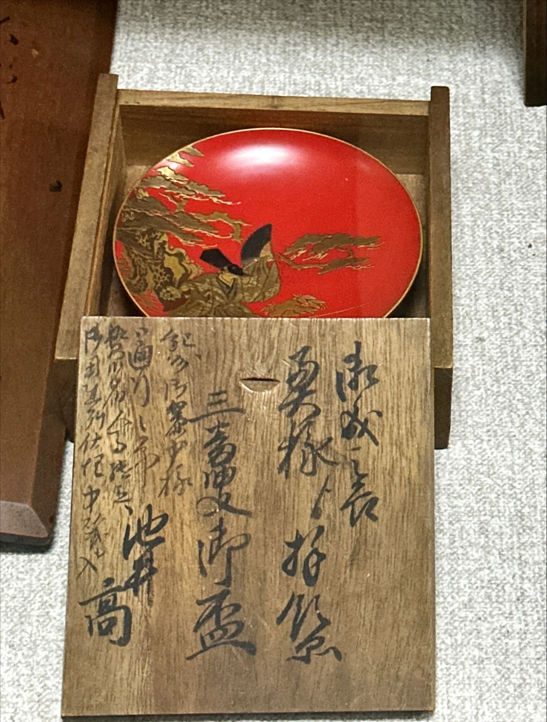 A red decorative urushi lacquerware antique plate set with gold accents and a black butterfly design, sitting inside a wooden tomobako box with Japanese calligraphy on the front, on a speckled surface.
