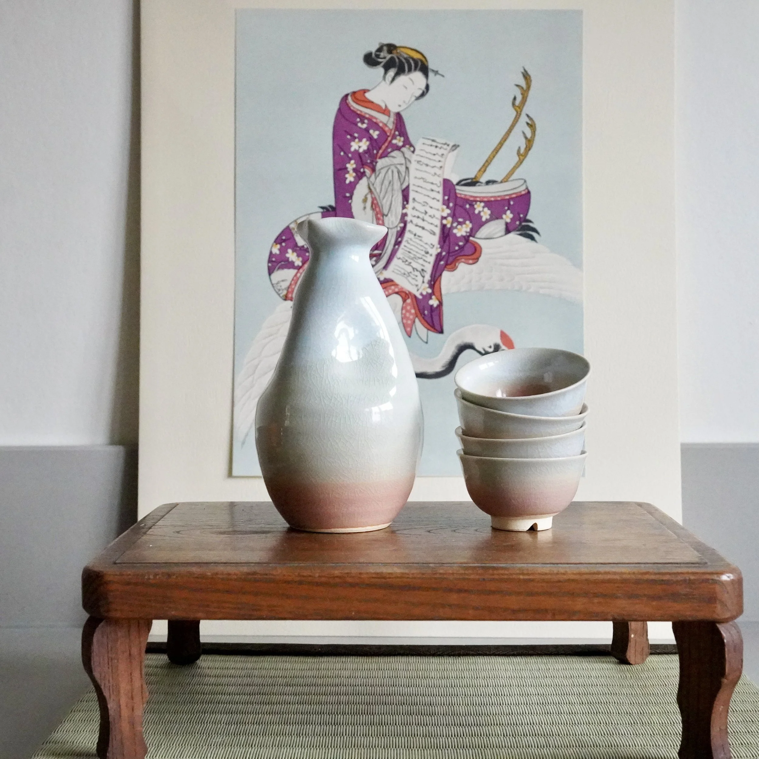 Tokkuri and four stacked ochoko with a pink to light blue crackled glaze, on a low wooden table. A tatami mat and Japanese art print are in the background.