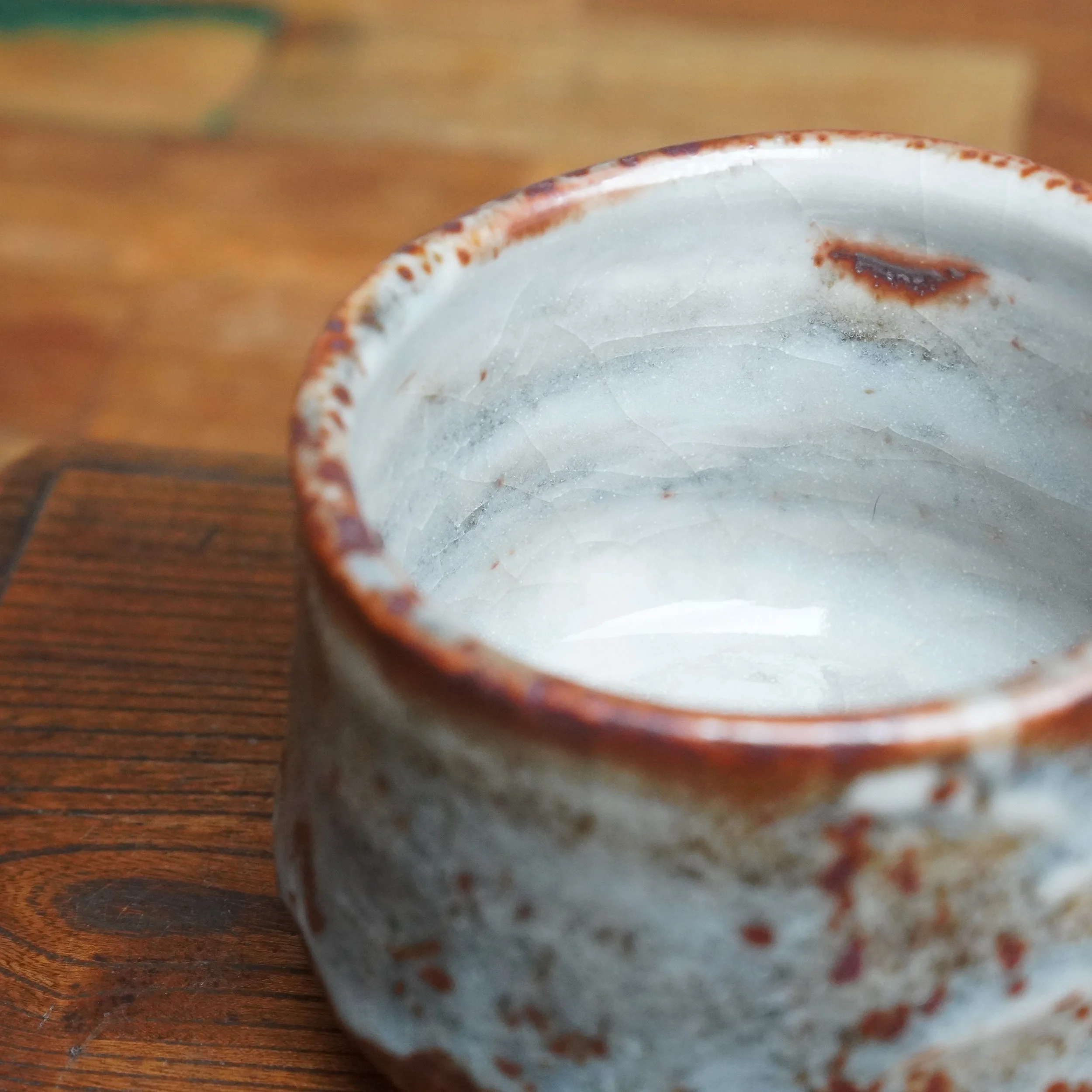 Shino winter chawan, angled view of its pale grey interior with a fine web of cracked glaze and deep red speckles, rimmed in rustic reddish-brown.