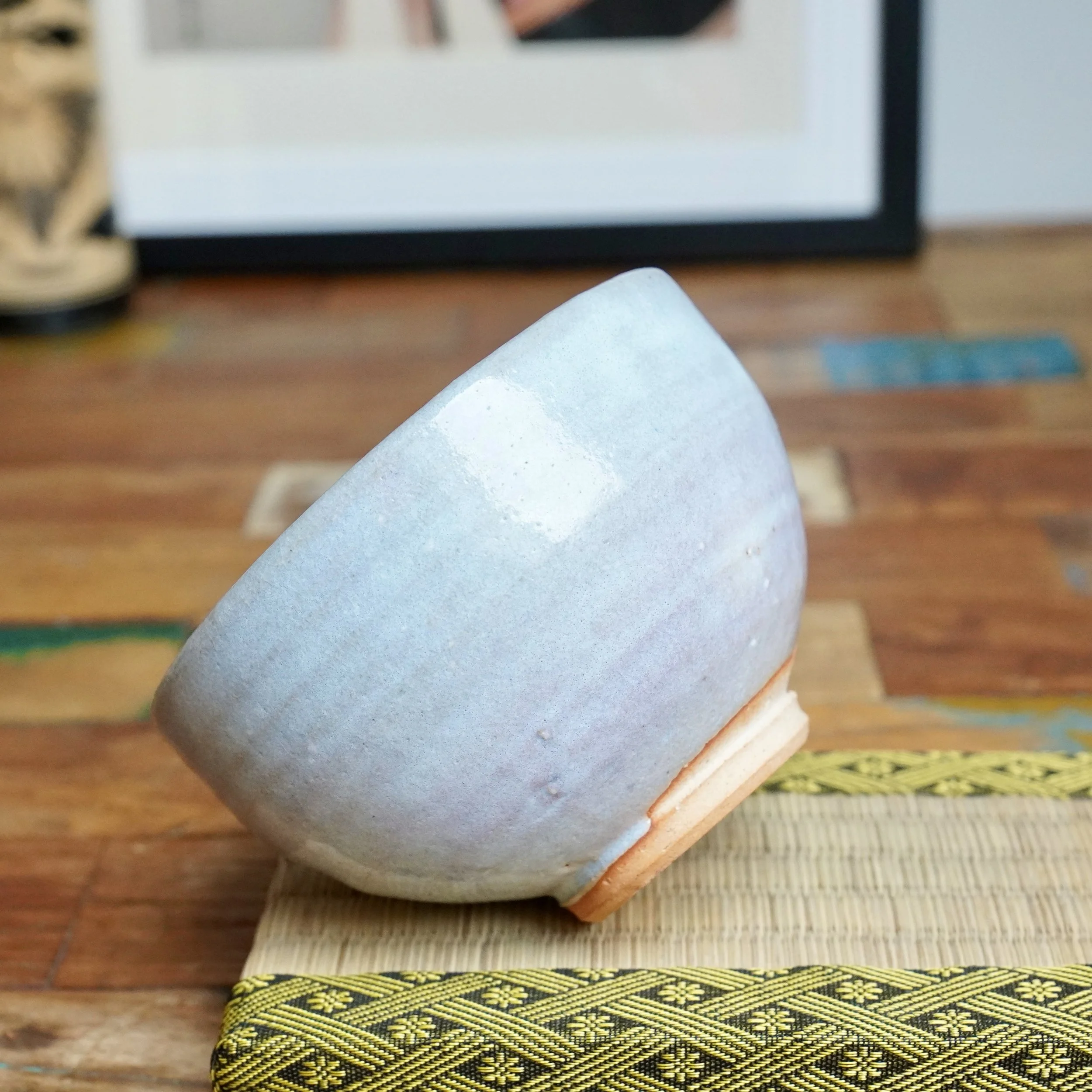 Moriwaki Fumitada chawan, tilted side view revealing soft white-blue glaze, unglazed terracotta foot, on a woven mat with yellow and black border.