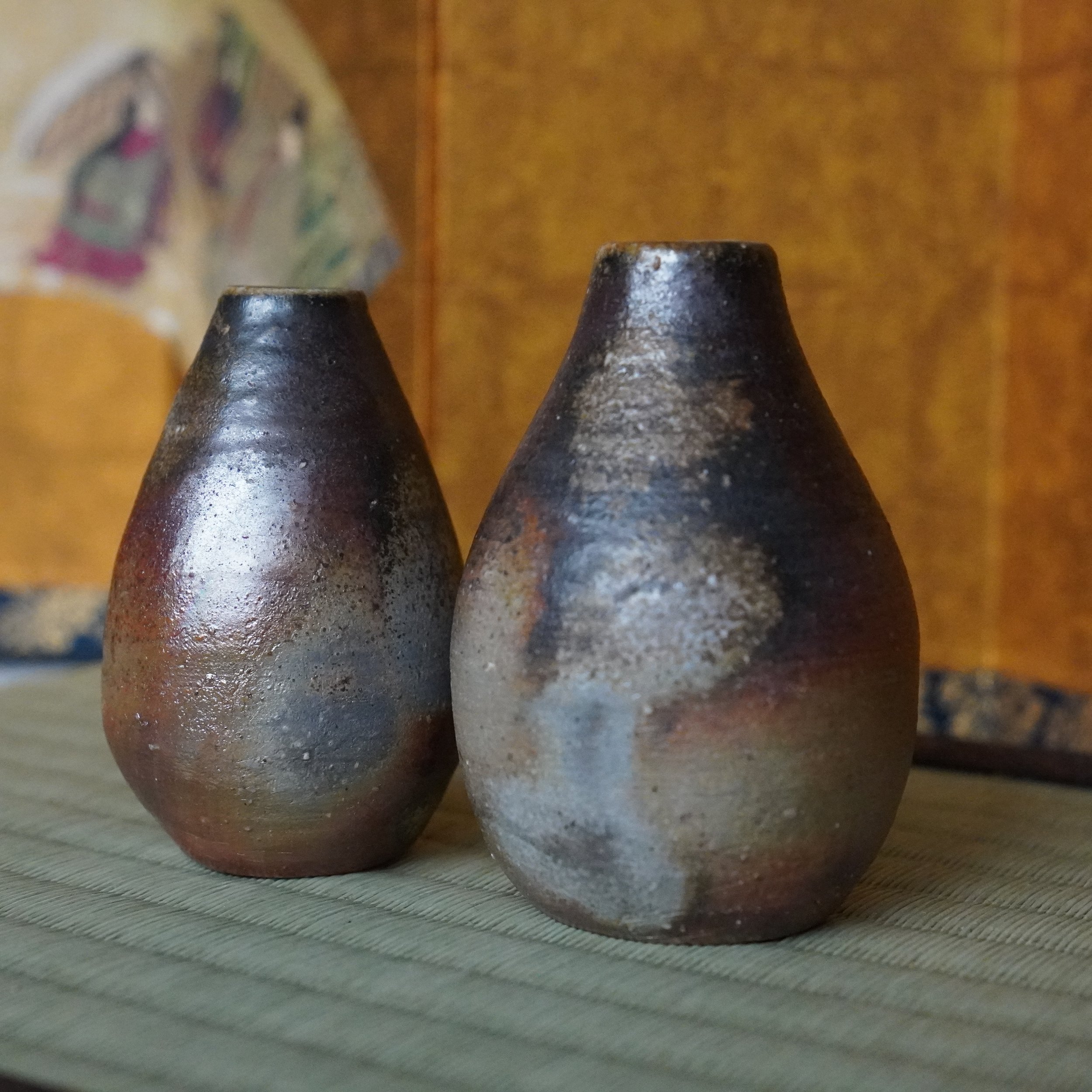 Shibaoka Tōsendō Bizen-yaki tokkuri pair: one aubergine and silver, one silvery-grey and rust. Placed on a tatami mat, with a golden blurred background.